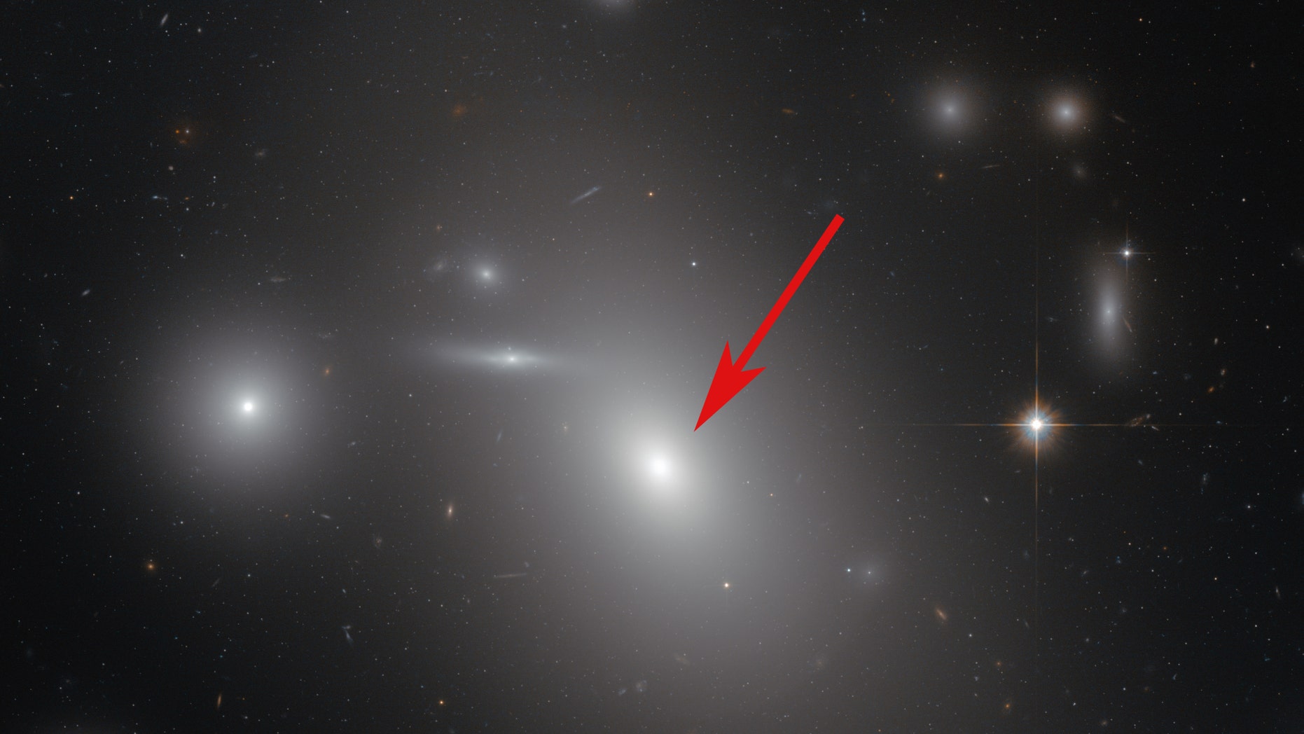 This image taken by the Hubble Space Telescope shows the elliptical galaxy NGC 4889 (arrowed) in front of hundreds of background galaxies. Well-hidden from human eyes, there is a gigantic supermassive black hole at the centre of the galaxy. (Credit: NASA & ESA)