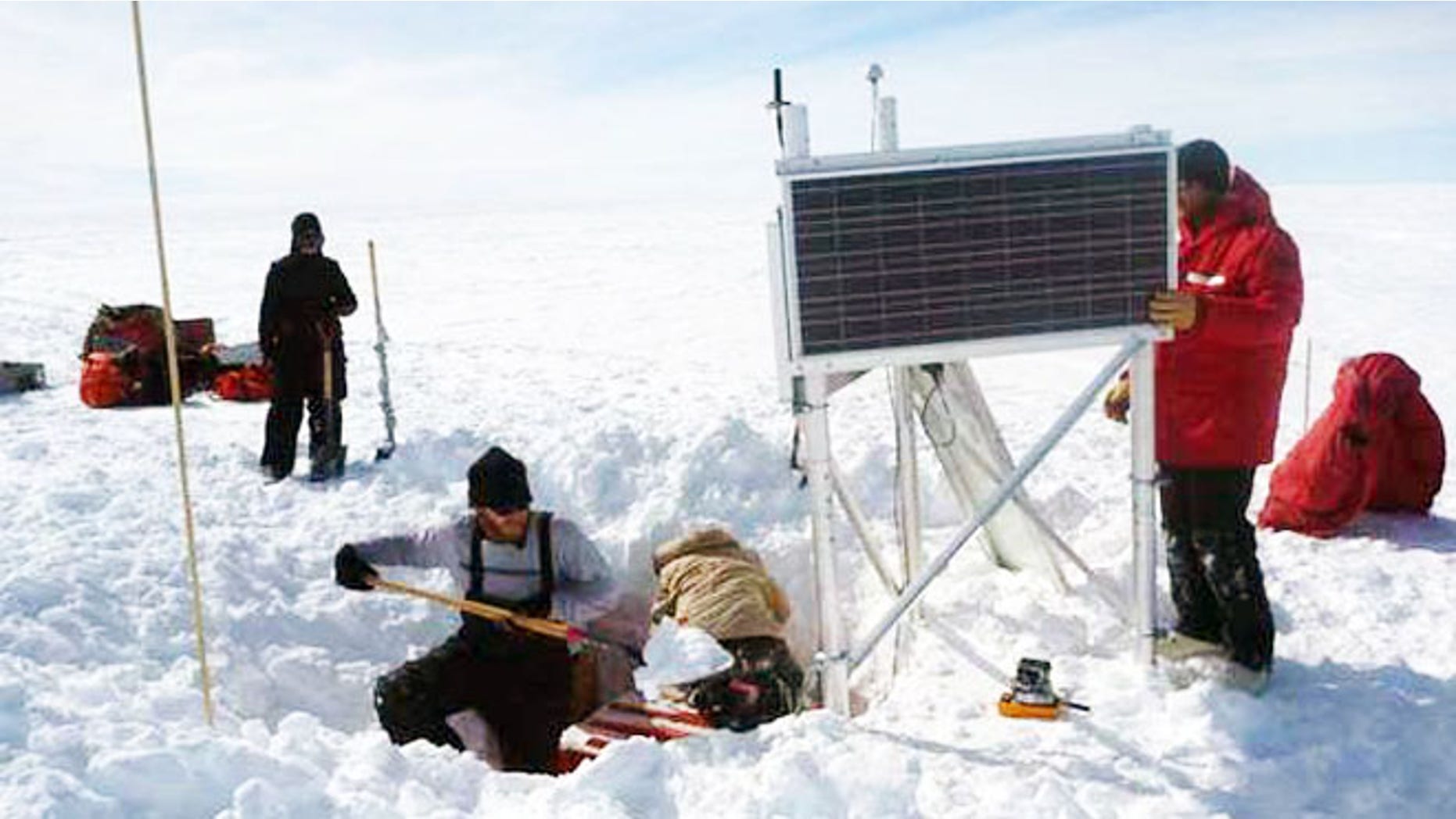 Researchers hard at work around a seismograph, an instrument in the orange box buried in a hole in the snow. Solar power runs the seismic station during the summer, and batteries keep it going during the long, dark winter months.