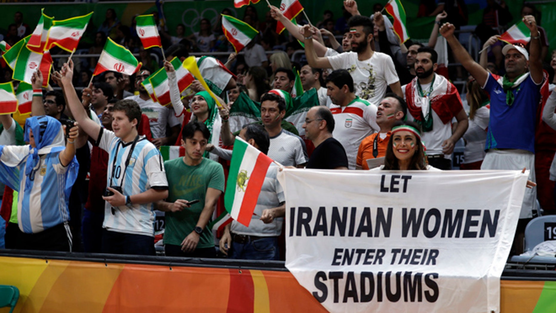 Fans cheer and wave the flag of Iran as Darya Safai, right, holds a large sign protesting the fact that women have not been allowed to attend volleyball matches in Iran during a men's preliminary volleyball match between Egypt and Iran at the 2016 Summer Olympics in Rio de Janeiro, Brazil, Saturday, Aug. 13, 2016.