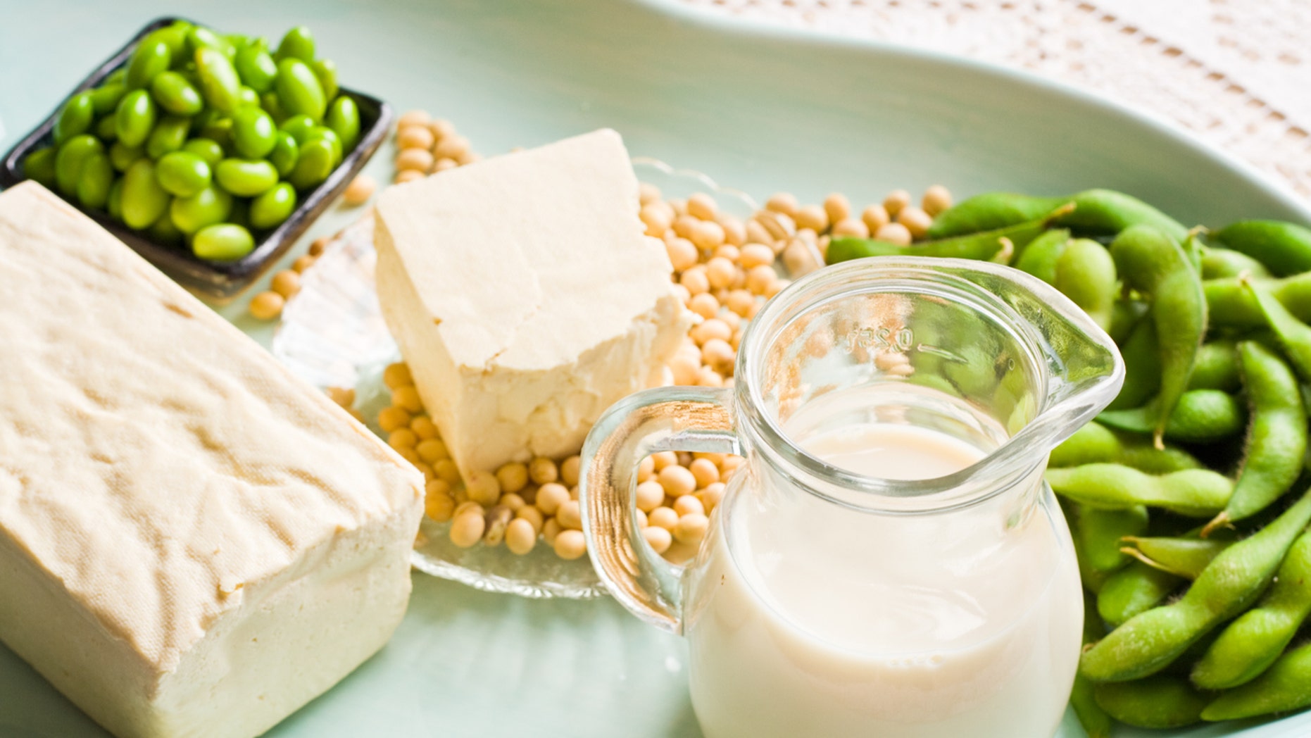 A variety of soy milk and soybean products. They are sitting in a soft aqua colored tray. There is a brick of firm tofu and a small pitcher of soy milk. On the right are some green soy beans and in back there is a small dish of shelled soybeans.