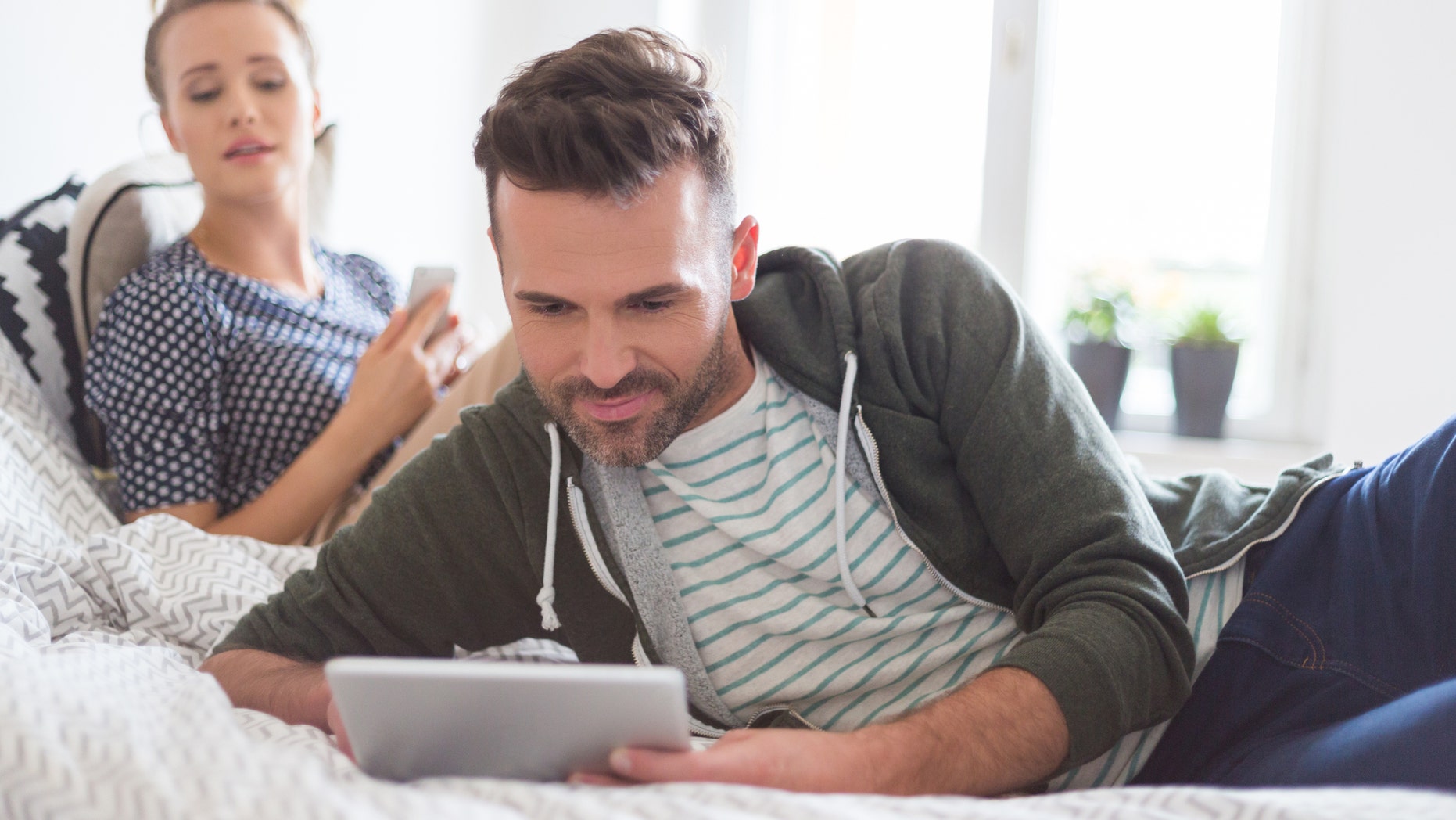 Man in casual outfit lying in the bed at home and using a digital tablet. His girlfriend in the background.