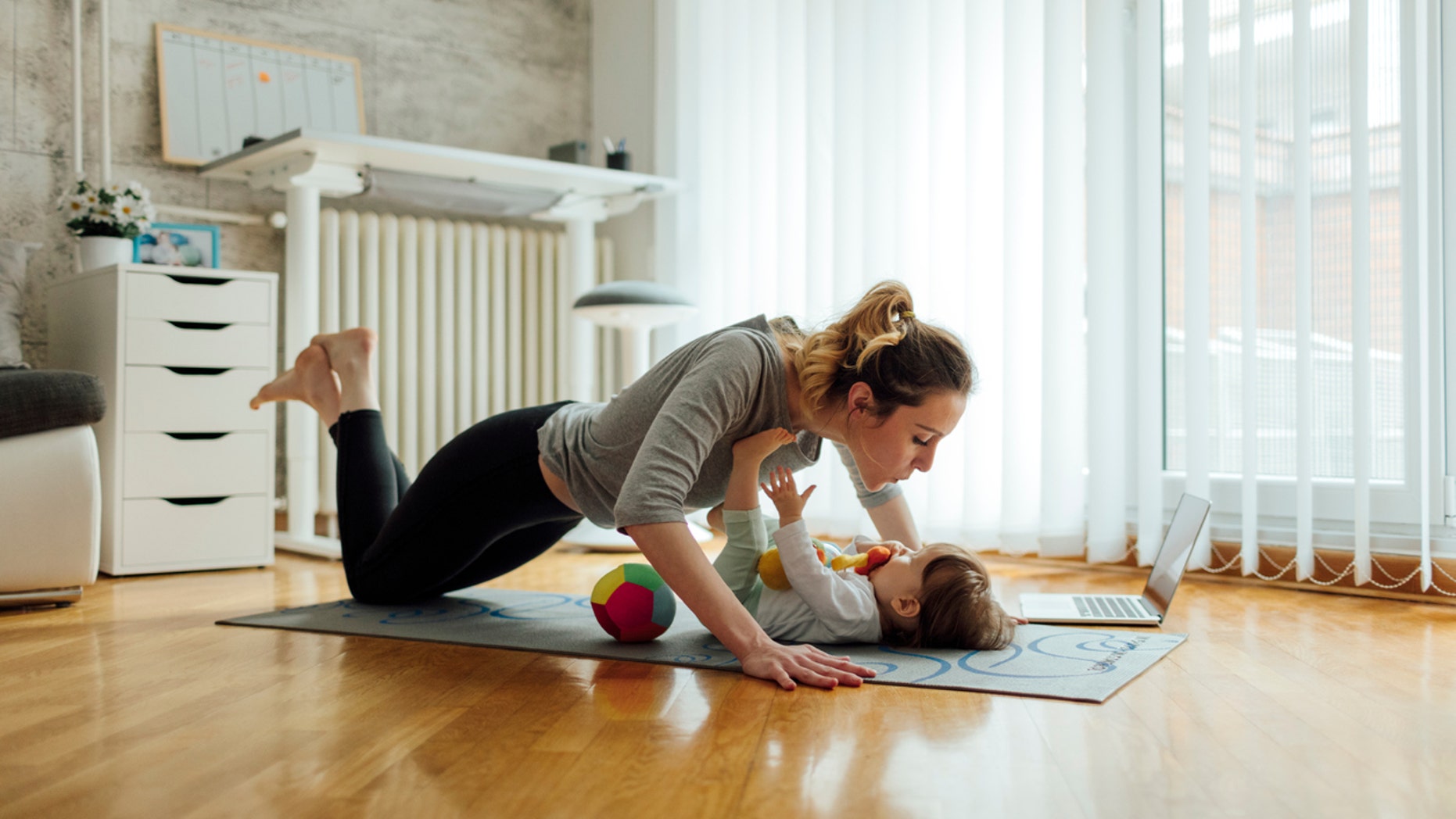 Shot of an attractive young woman with her baby girl while doing exercise. Woman working pushups and baby lying on exercise mat. They exercise in a living room. Woman watching exercise on laptop.