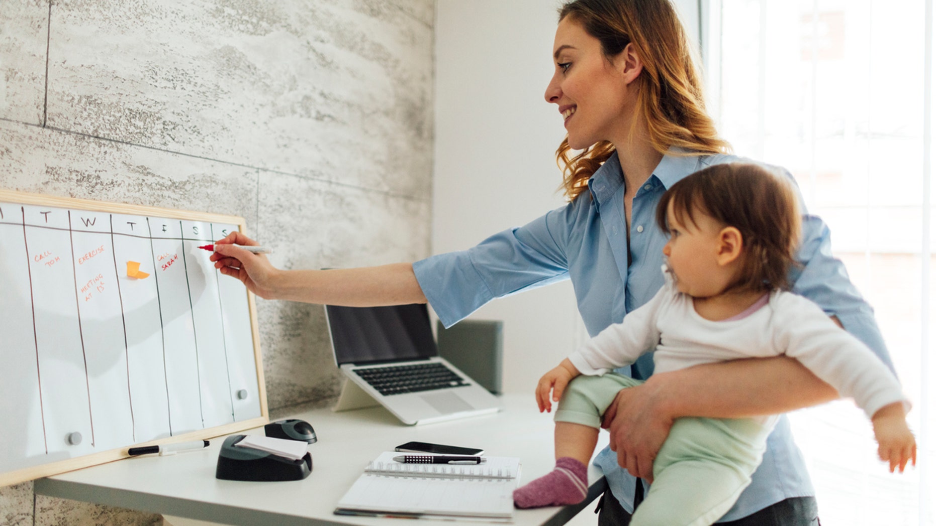 Mother working from home while carrying her cute baby girl. She is writing on whiteboard, entering into the schedule. She is happy and cheerful.