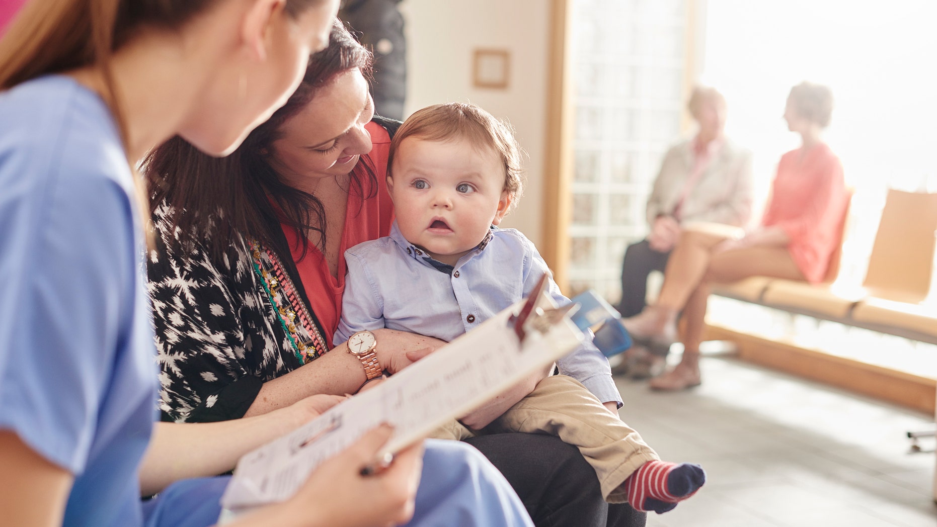 a female nurse sits next to a woman and her baby son in the waiting room of a clinic and casually chats to them . In the background two patients sit and chat to a senior doctor .