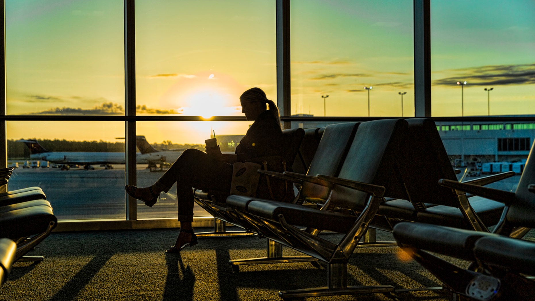 A woman sits at the airport waiting for her plane. She is enjoying a drink and snack as the sun rises.