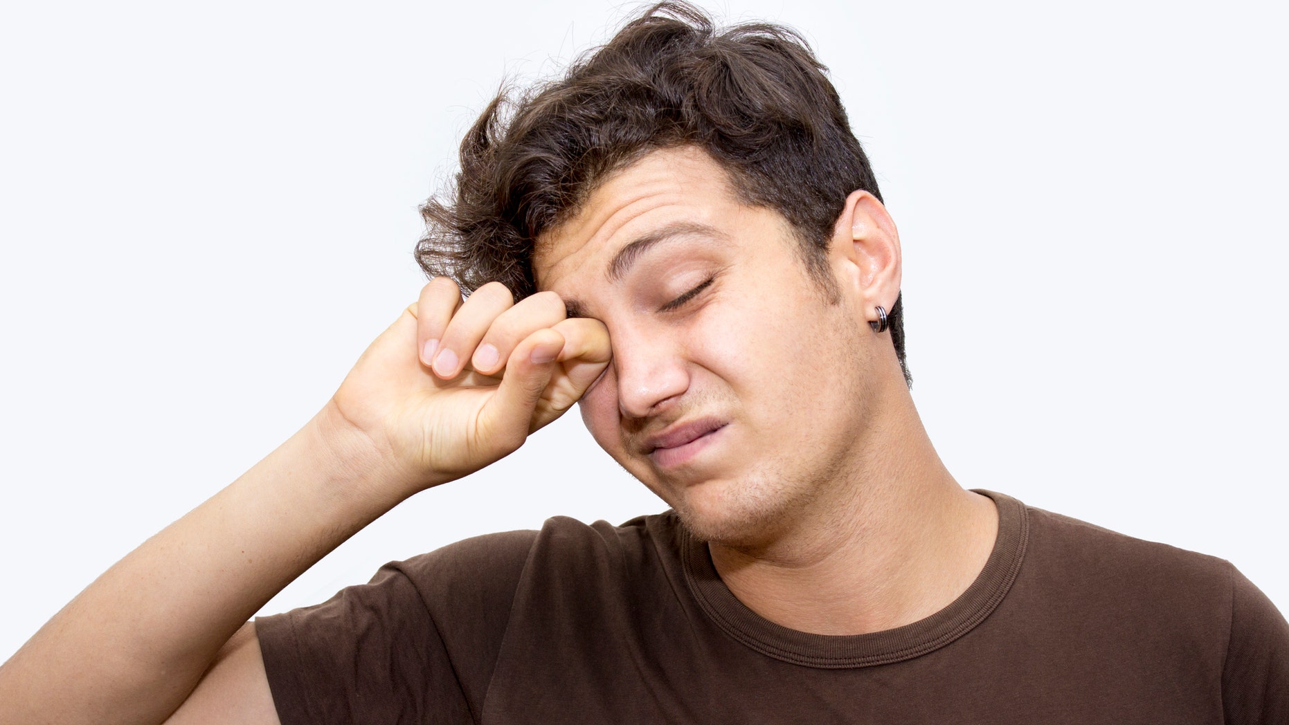 Portrait of napping young man rubbing his eyes isolated on white. Horizontal composition. Studio shot. Image developed from RAW format. Tired teenage boy has got short, curly, brown hair.