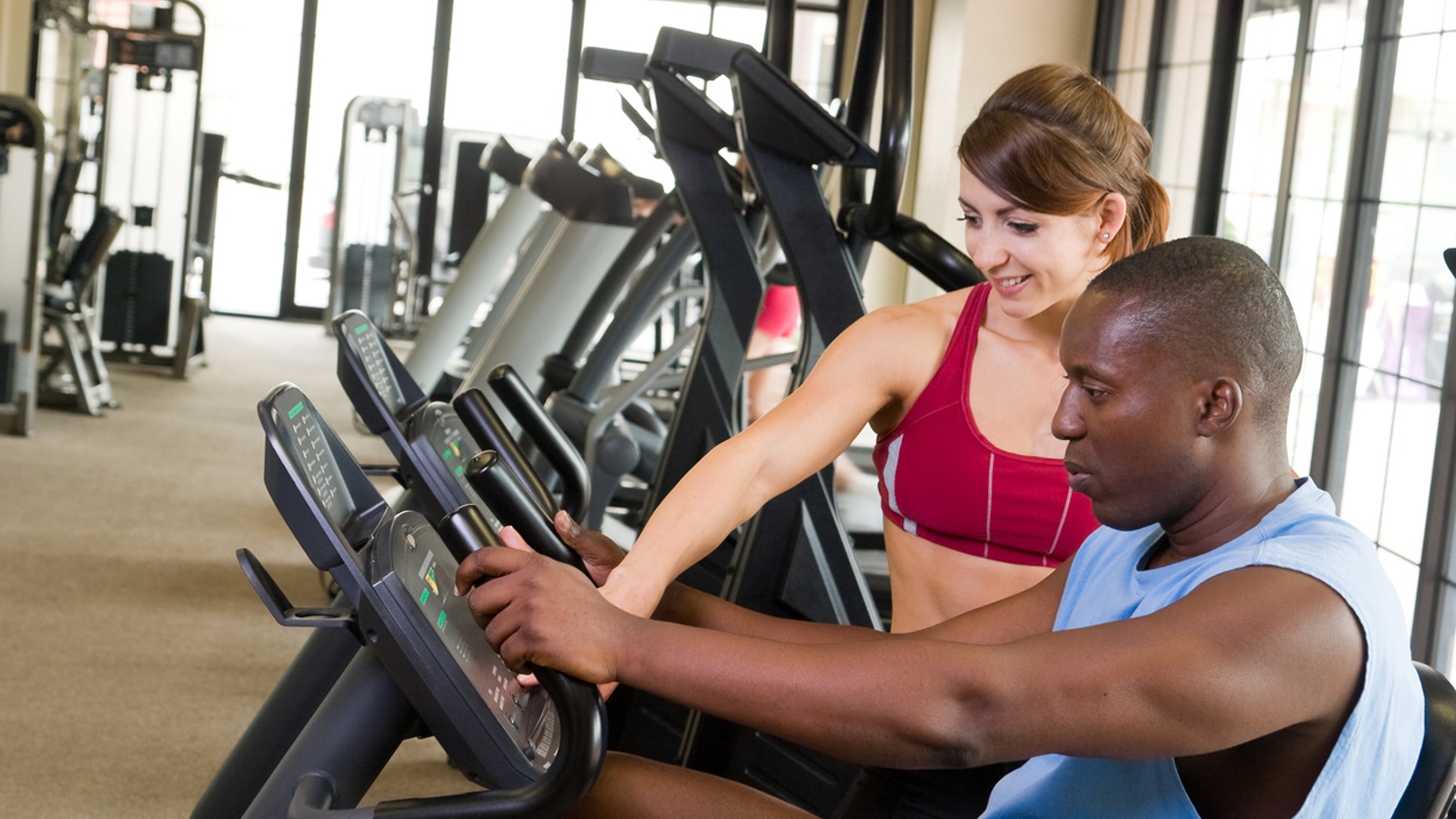 Man and woman exercising together at a fitness center on a stationary bicycle exercise machine. Woman could be a personal fitness trainer. Focus is on the man in the foreground.
