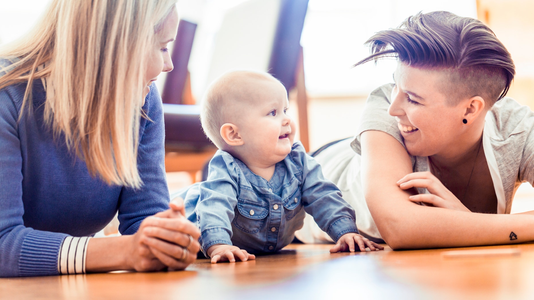 Smiling lesbian couple with baby lying on hardwood floor. Homosexual partner with toddler in living room. Happy family of three at home.