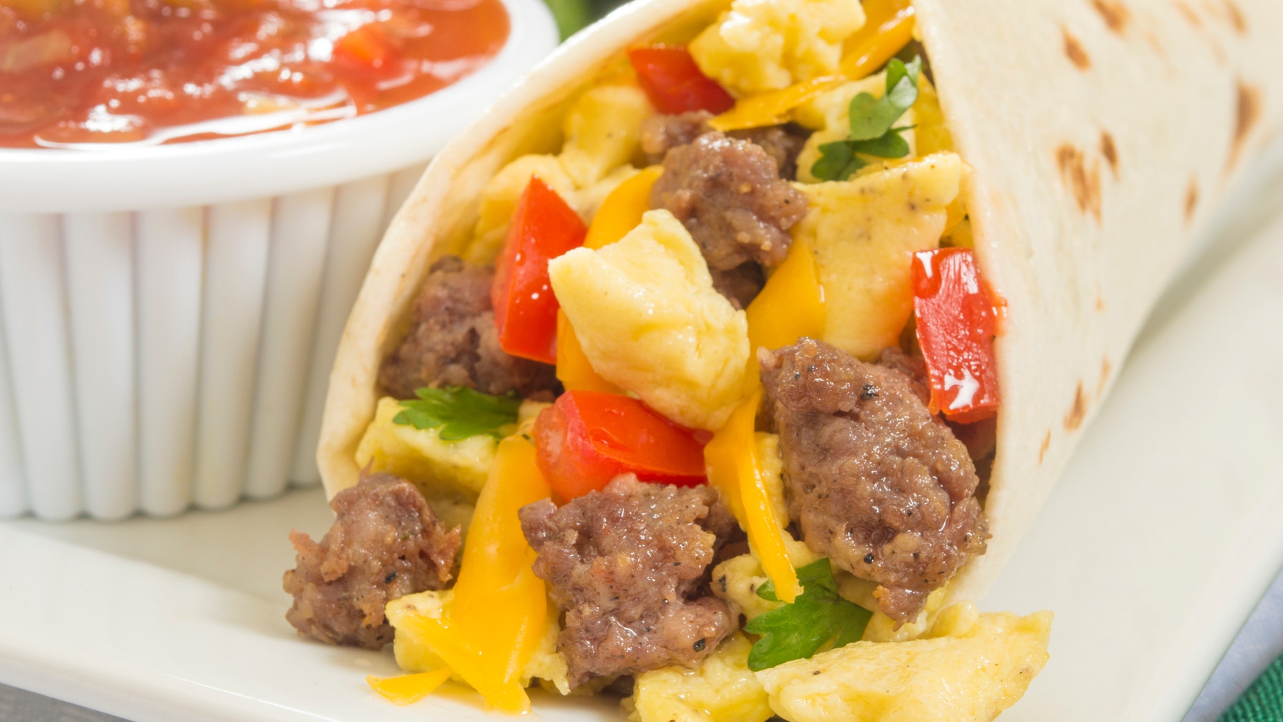 close up of a hot fresh breakfast burrito with eggs,sausage,cheese,tomatoes,cilantro, and salsa on the side