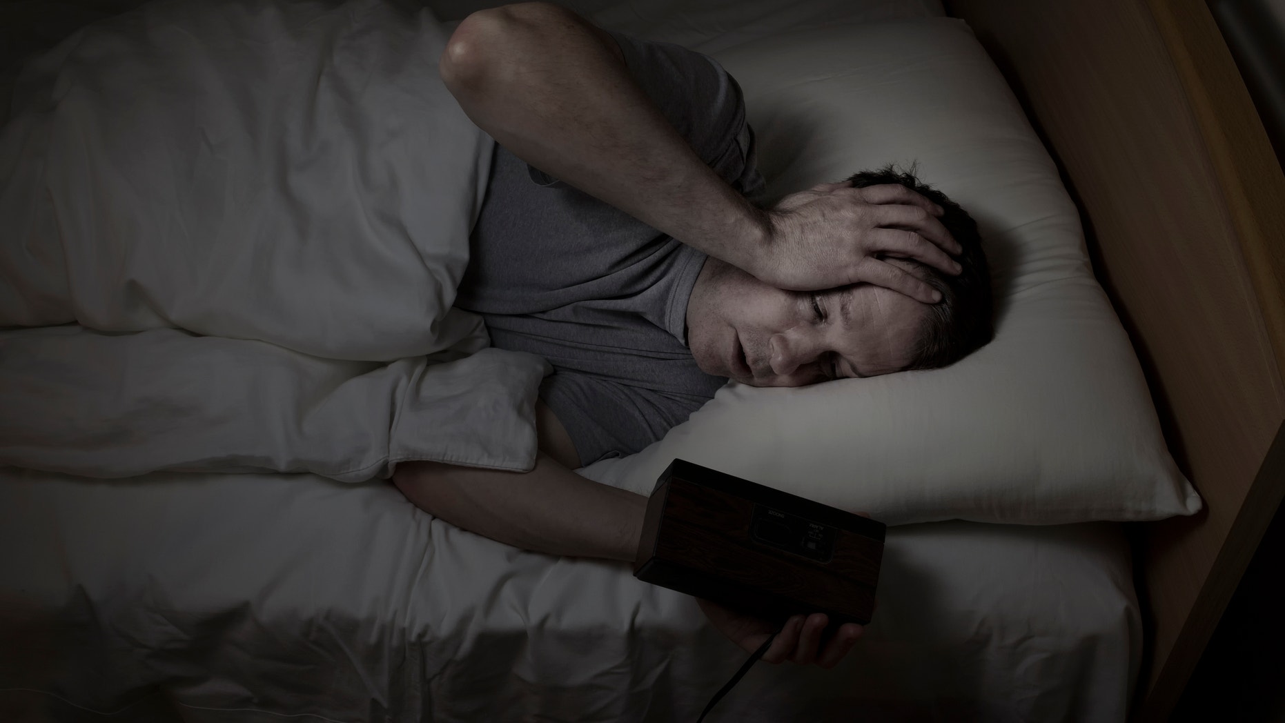 Mature man, eyes open with one hand holding clock and the other his own forehead, cannot sleep at night from insomnia