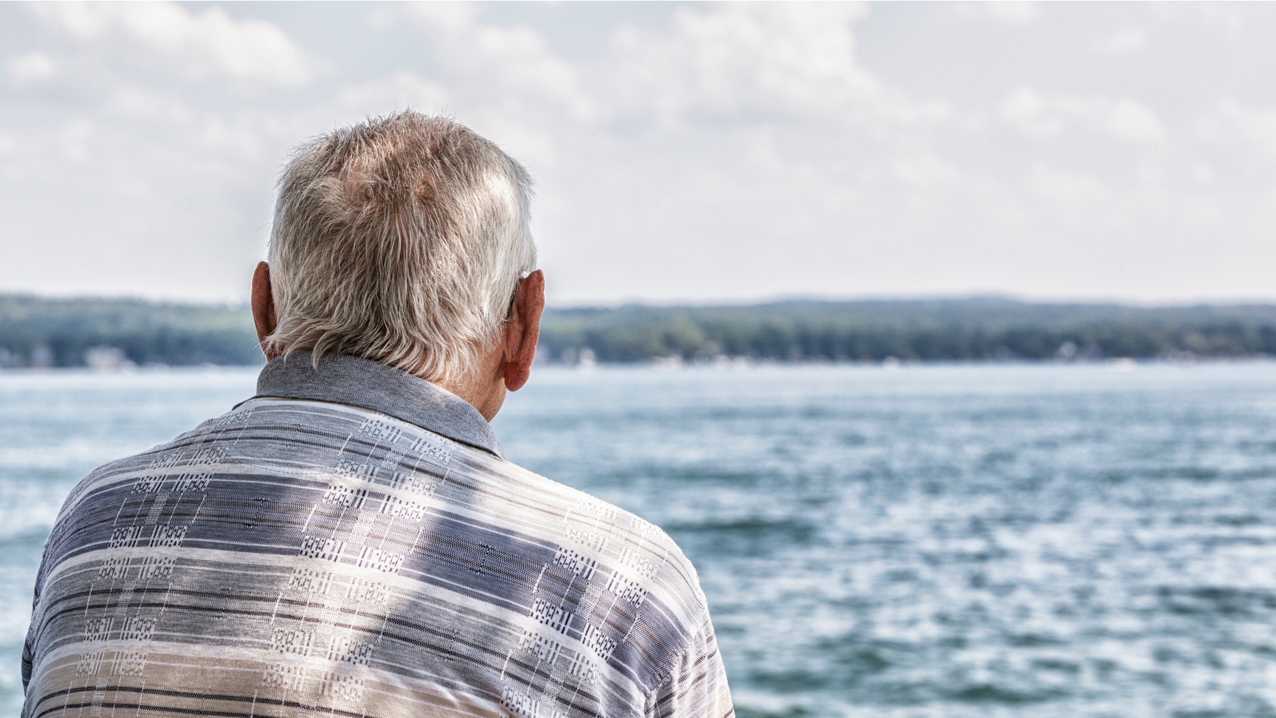 Rear view of a solitary elderly 80 plus year old senior adult man sitting at the edge of the lake watching the boats and other recreation activity on the water.