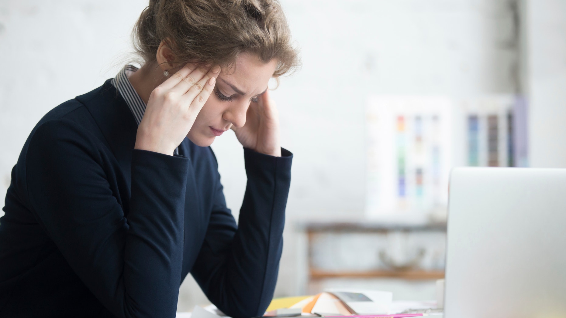 Portrait of young stressed woman sitting at home office desk in front of laptop, touching head with frustrated facial expression, having headache, overworked or depressed