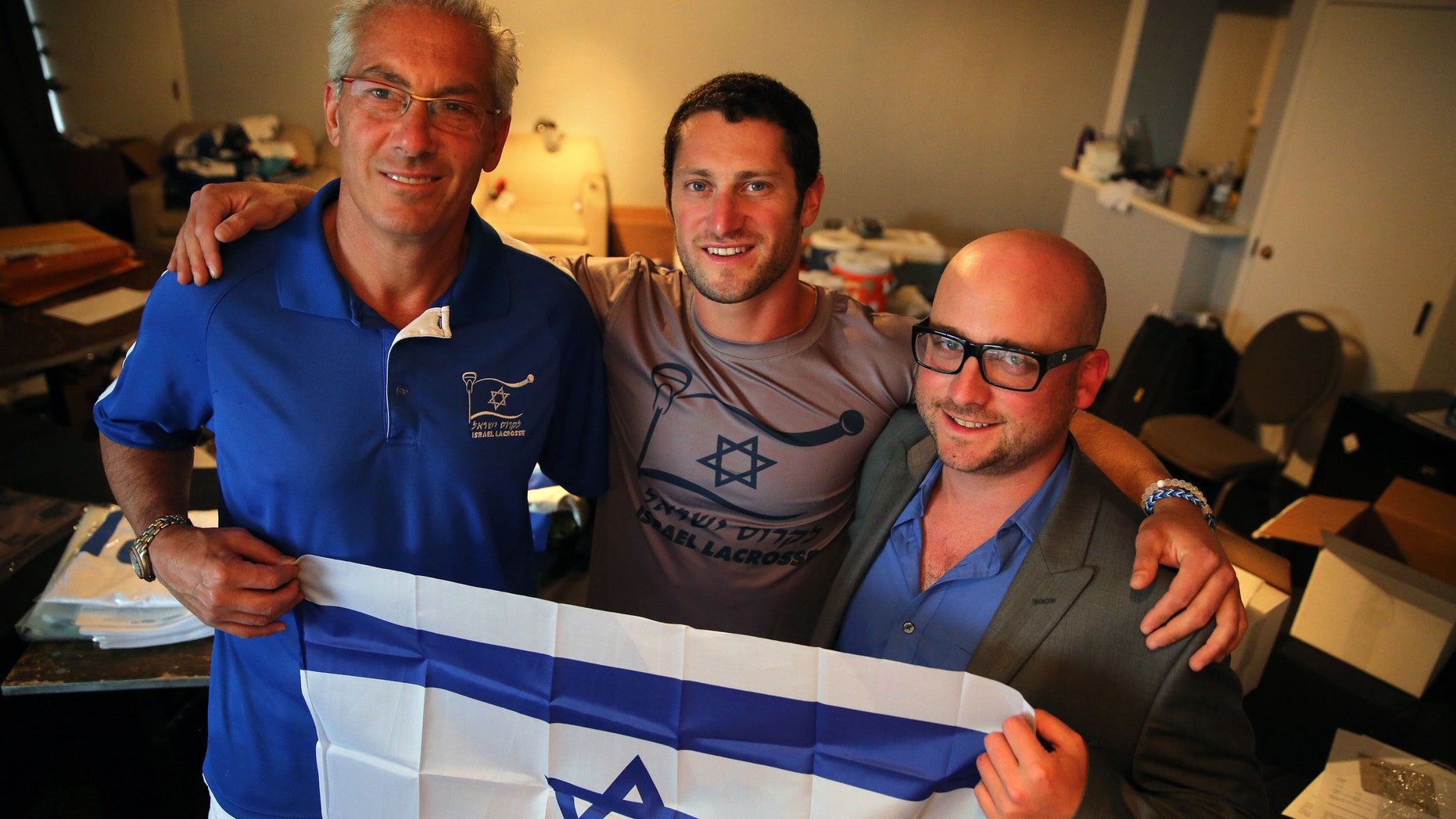 July 15, 2014: In this Israel Lacrosse head coach Bill Beroza, left, attackman Noach Milller, center, and team executive director Scott Neiss hold an Israeli flag inside a hotel room used to organize team gear, in Commerce City, Colo.