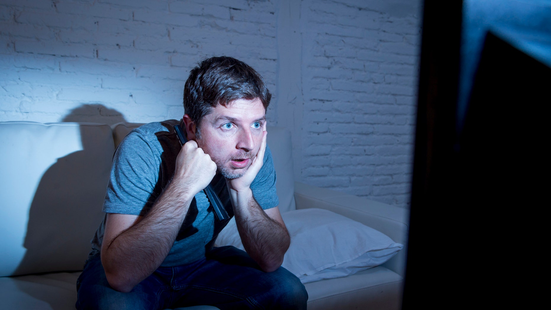 young attractive man at home lying on couch at living room watching tv holding remote control looking mesmerized and intense in television addict concept