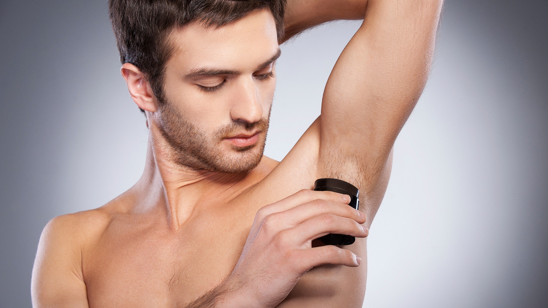 Staying fresh. Confident young shirtless man using dry deodorant while standing isolated on grey background