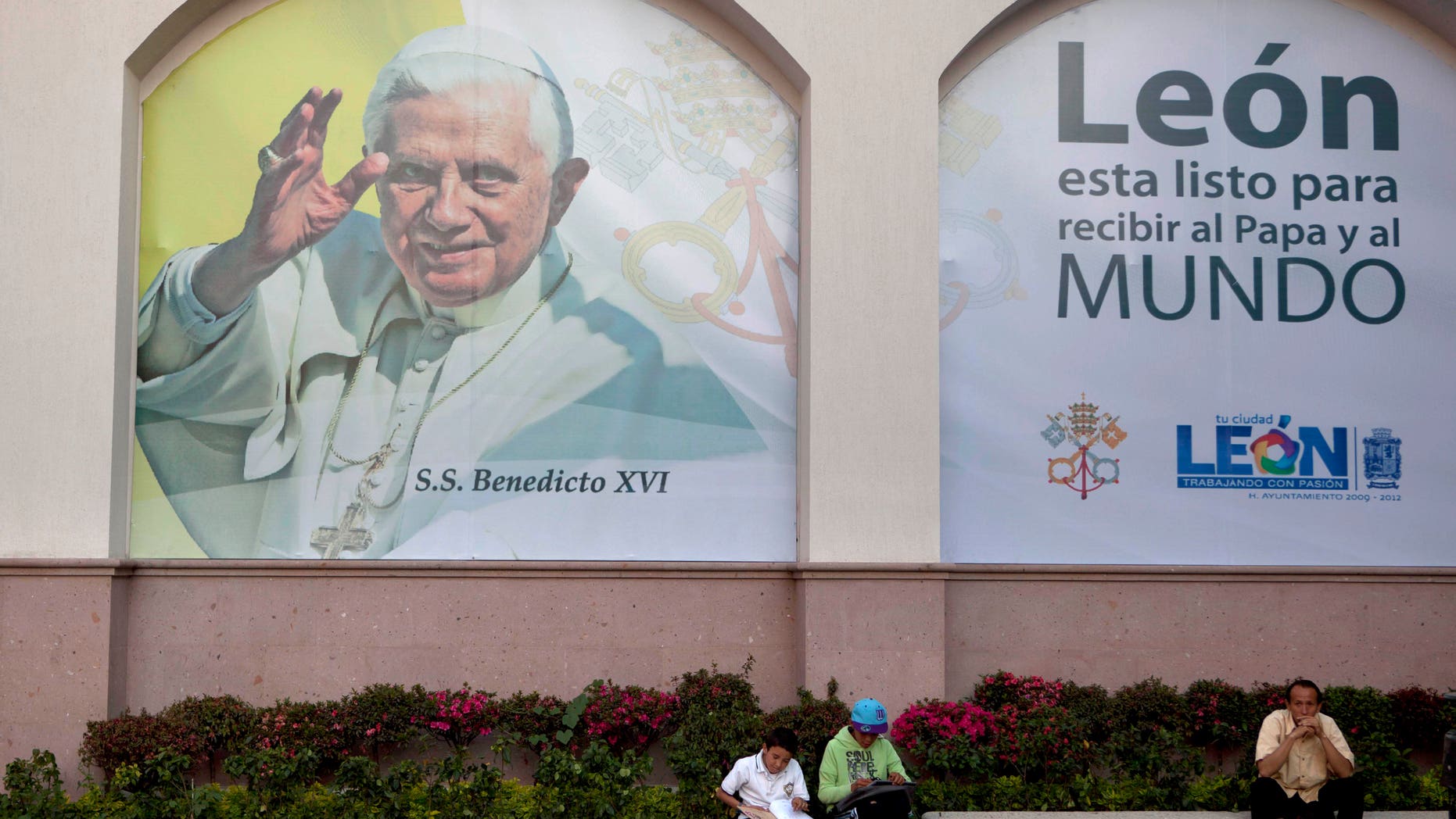 People sit in front of a banner placed over a wall with an image of Pope Benedict XVI that reads in Spanish "Leon is ready to receive the Pope and the world," in Leon, Mexico, Tuesday March 20, 2012. Benedict XVI will visit Mexico for the first time on March 23. (AP Photo/Eduardo Verdugo)