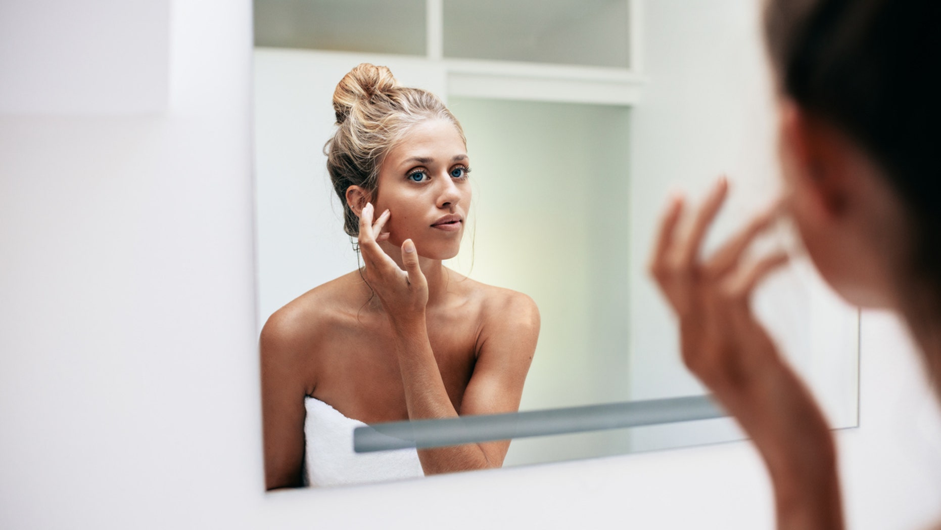 Beautiful young woman standing in the bathroom. Female looking into the mirror and touching her face skin.