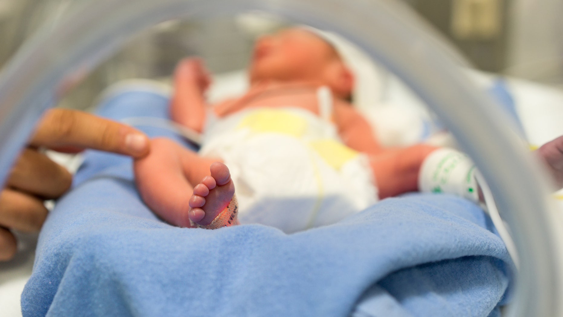 Photo of a premature baby in incubator. Focus is on his feet and toes. The doctor is touching him to check his reflexes. There are cables and tubes in the out-of-focus area.