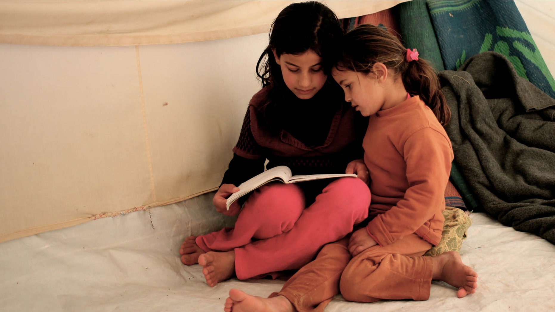 Syrian refugee girls, Fatima Teeme, right, and Sara Teeme, from Homs, read a book inside their tent, as their father Ibraheem Teeme speaks to The Associated Press, at Zaatari refugee camp, near the Syrian border in Mafraq, Jordan, Monday, Dec. 16, 2013. NRC began distributing stoves to Syrian refugees at the Zaatari camp in Jordan on Monday, as bitter winter weather made living conditions worse. (AP Photo/Mohammad Hannon)