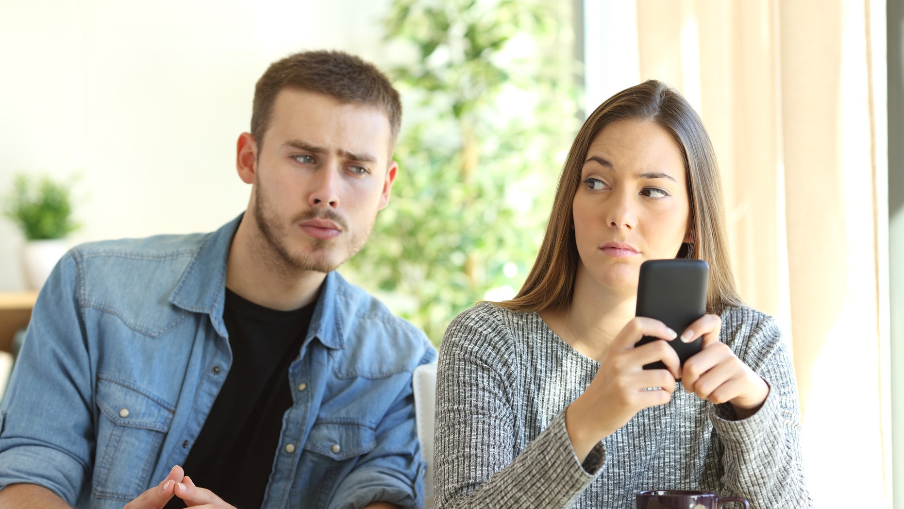 Jealous boyfriend spying his girlfriend watching her phone while she is looking him upset (Credit: AntonioGuillem, iStock)