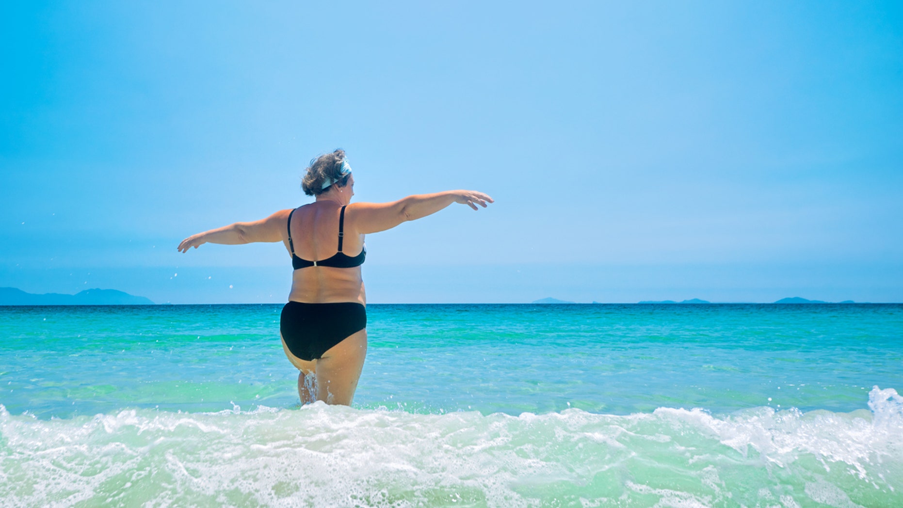 mature woman in swimwear at the sea. Overweight woman in swimsuit comes in water in time of surf, rear view