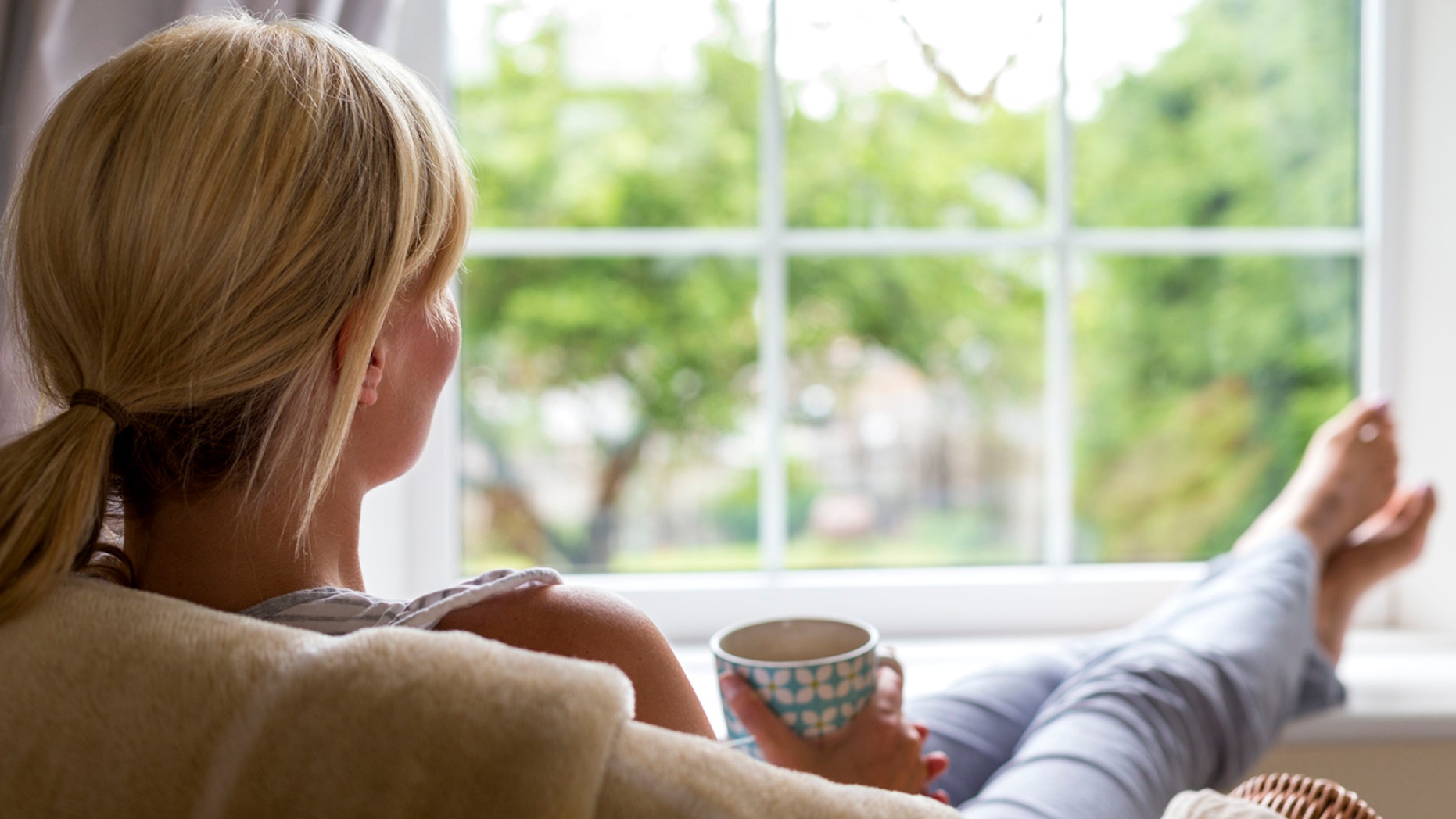 A young woman sits with a hot drink and her feet up, watching out of the window from the comfort of her own home. She is taking a break from her chores and busy day to day routine to gaze into her garden and wonder.
