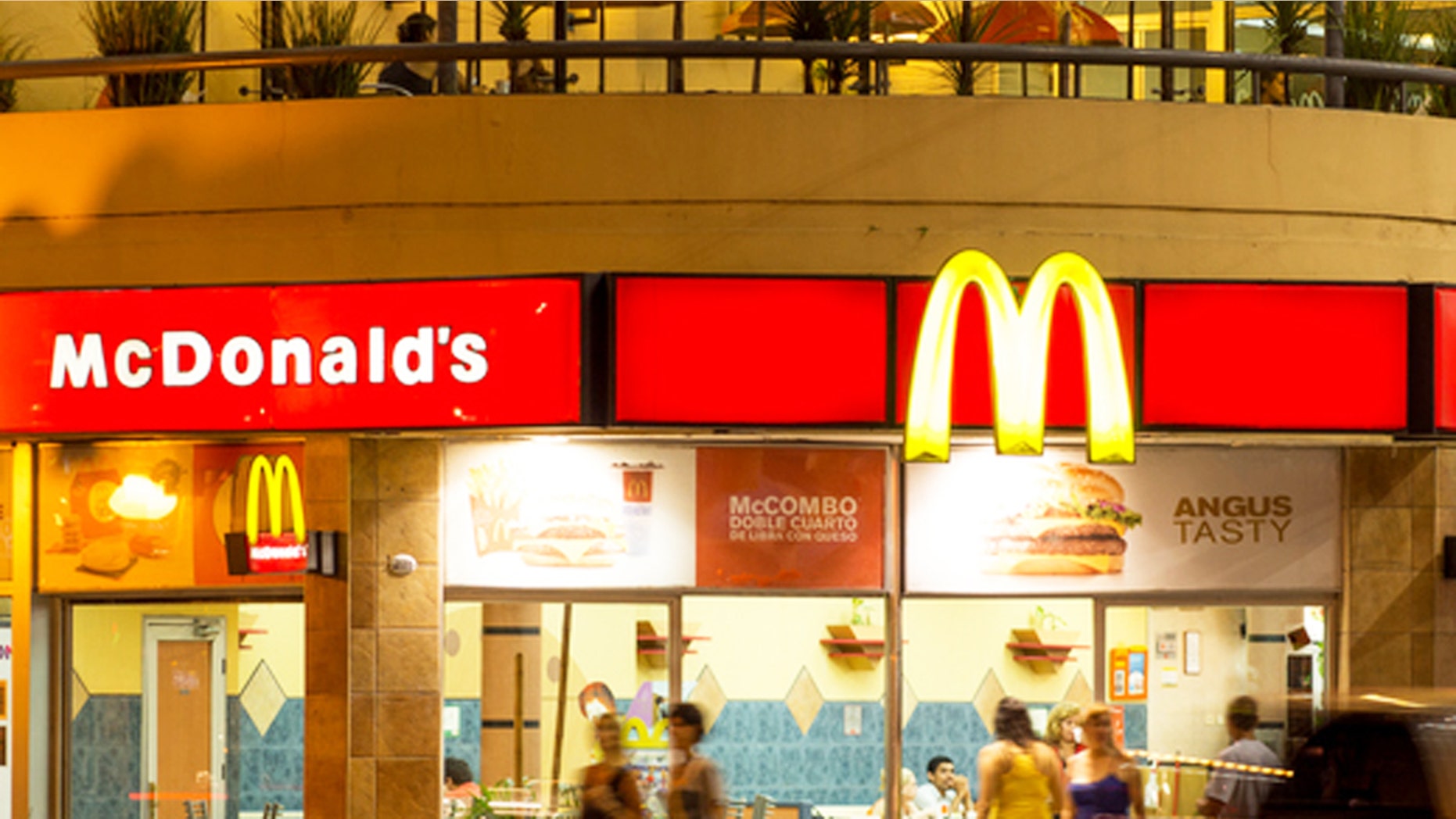 "Palermo, Buenos Aires, Argentina - January 22, 2013: Nighttime view of a McDonald's Restaurant with the light trail of a departing car in the foreground. People can be seen eating inside of the restaurant, entering the restaurant, and walking by on the sidewalk."