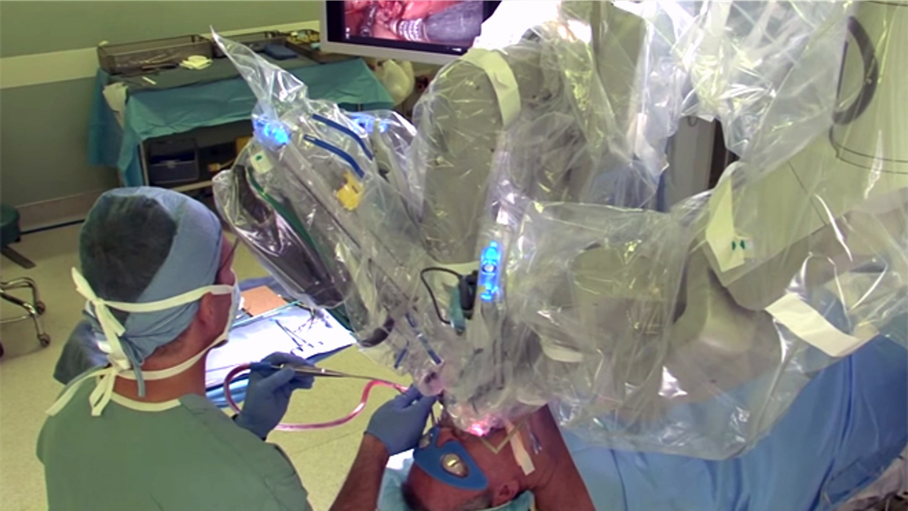 A physician monitors a patient undergoing Trans Oral Robotic Surgery (TORS) which is utilizing the Da Vinci robotic surgical system, and is being controlled by a surgeon who is not pictured.