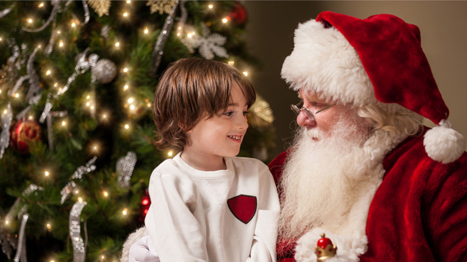 A young boy sits on Santa Claus' lap by a decorated Christmas tree in his living room. Santa has a real beard and the child looks at him with wonder, awe, and a hint of excitement.