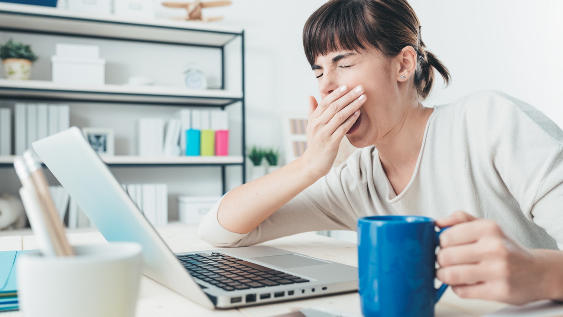 Tired sleepy woman yawning, working at office desk and holding a cup of coffee, overwork and sleep deprivation concept