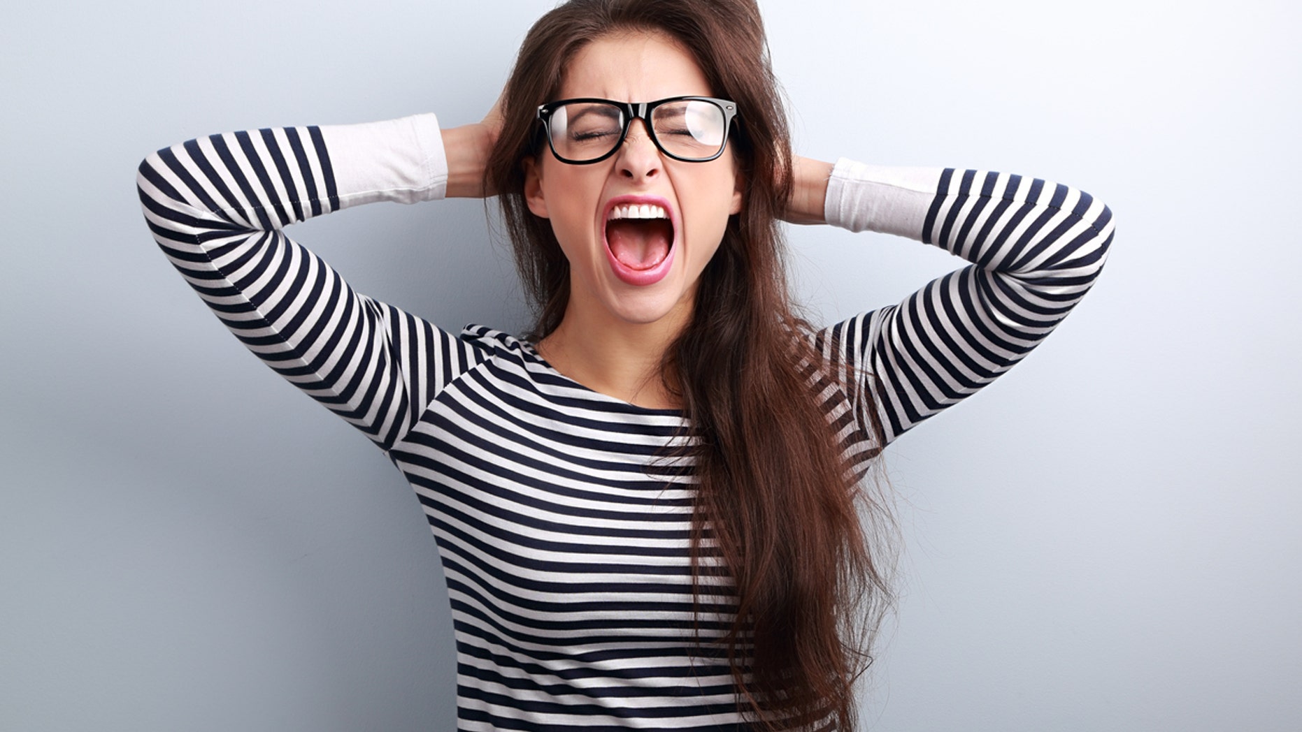 Anger young business woman in glasses strong screaming with wild open mouth and holding head the hands on blue background