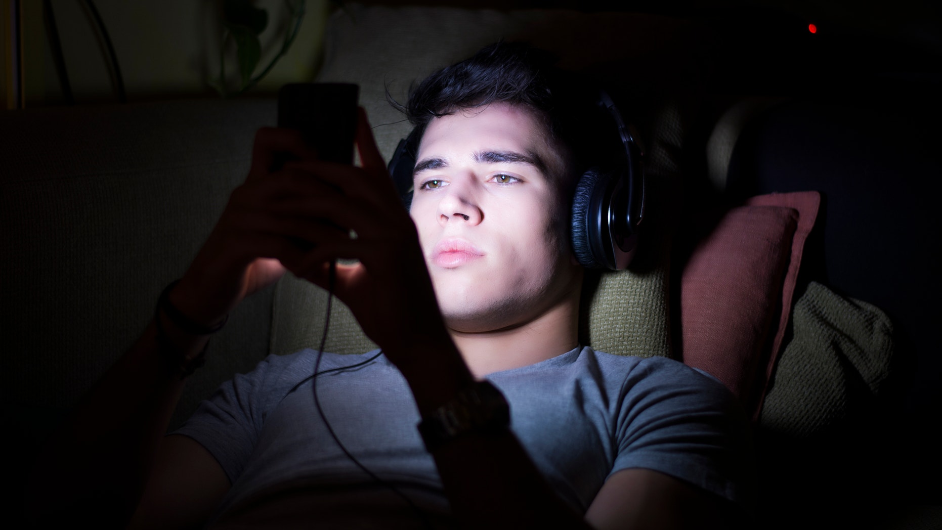 Attractive Young Man Lying on Sofa at Night and Illuminated by Light from Screen of Cell Phone at Home in Living Room