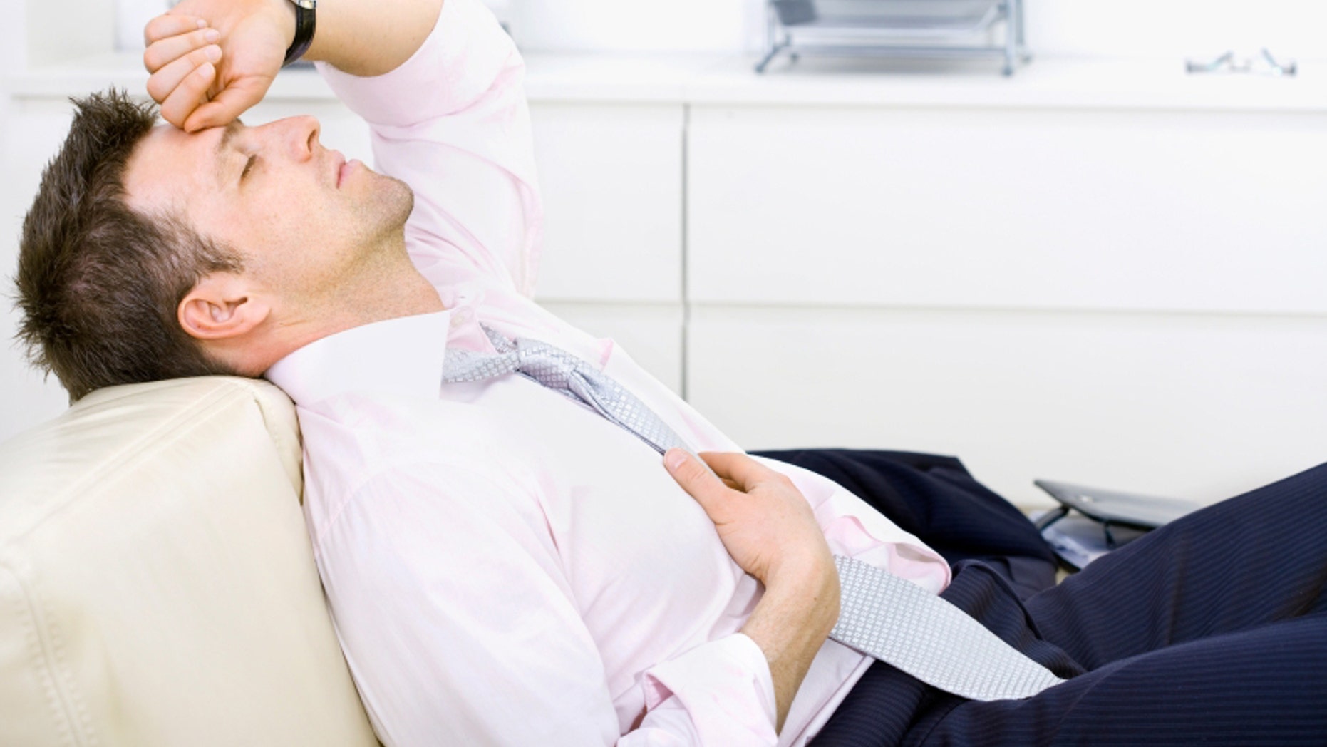 Mid-adult businessman lying on sofa at office, looking tired, eyes closed. Bright background.