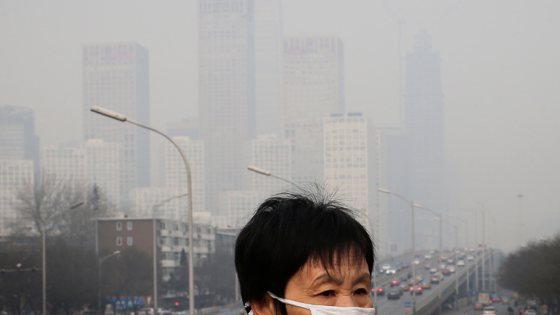 In this Dec. 20, 2015 file photo, a woman wearing a mask for protection against pollution walks on a pedestrian overhead bridge as office buildings are shrouded with smog in Beijing.