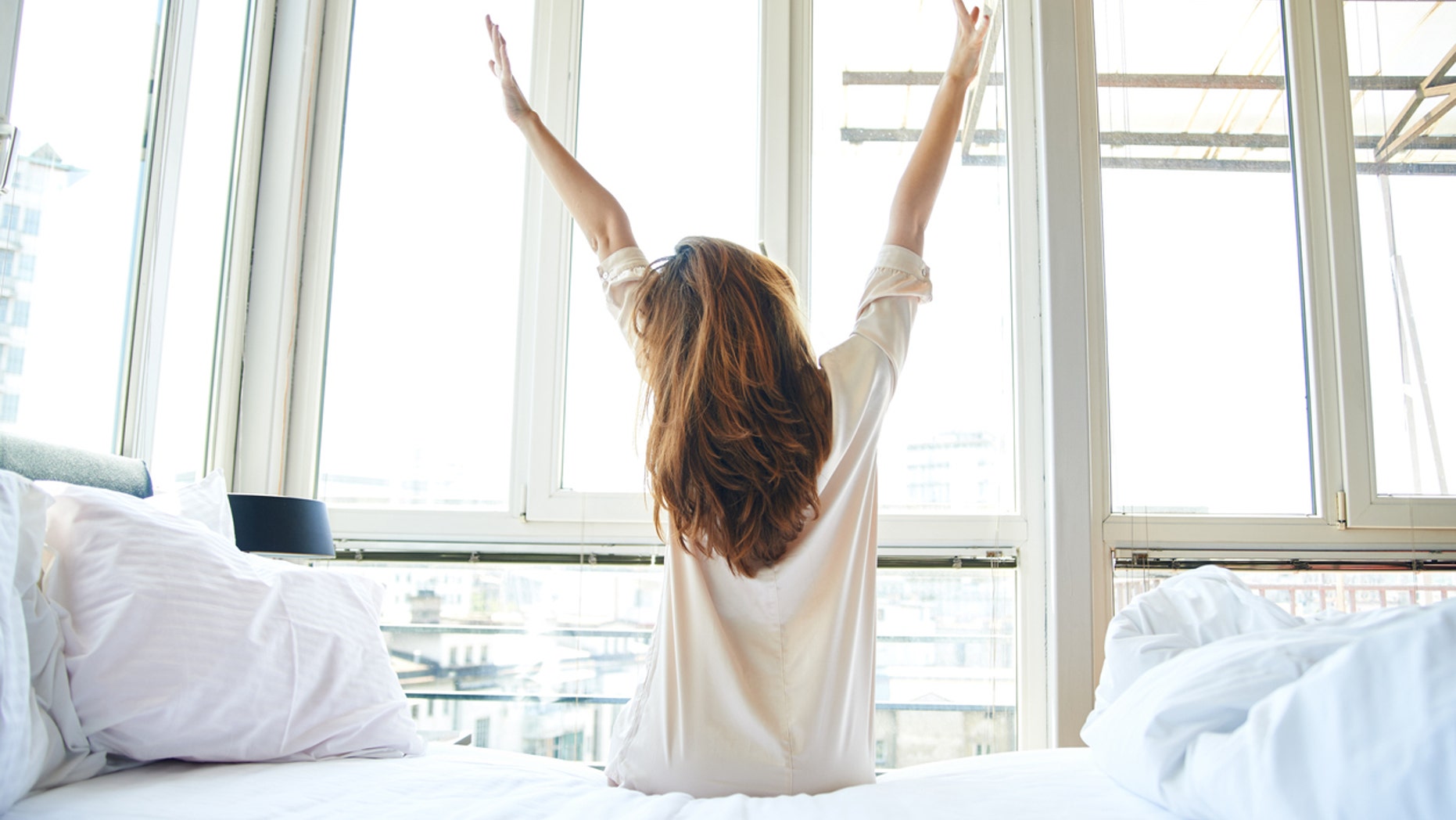 Young woman is doing morning stretching in bed, arms raised, rear view