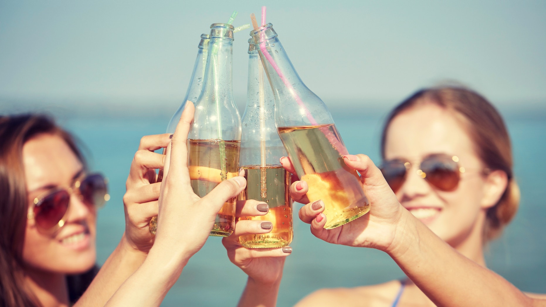 summer vacation, holidays, party, travel and people concept - close up of happy young women with drinks clinking bottles on beach
