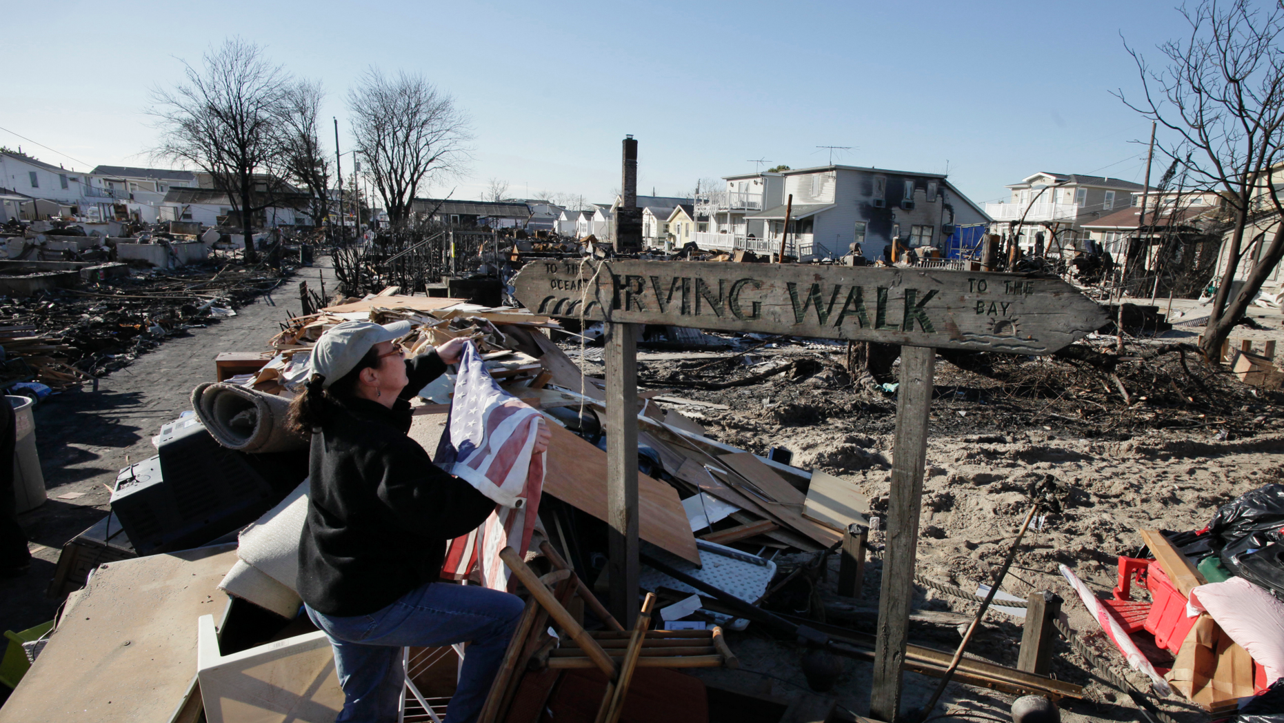 In this Nov. 14, 2012 photo, Louise McCarthy places an American flag on a street sign for Irving Walk in the Breezy Point neighborhood of Queens, N.Y. The sign survived a fire that swept through the seaside community during Superstorm Sandy two weeks earlier. Six years later, most residents have stayed. Homes have been restored, and Sandy led to a new ferry service and a rebuilt boardwalk. (AP Photo/Mark Lennihan)