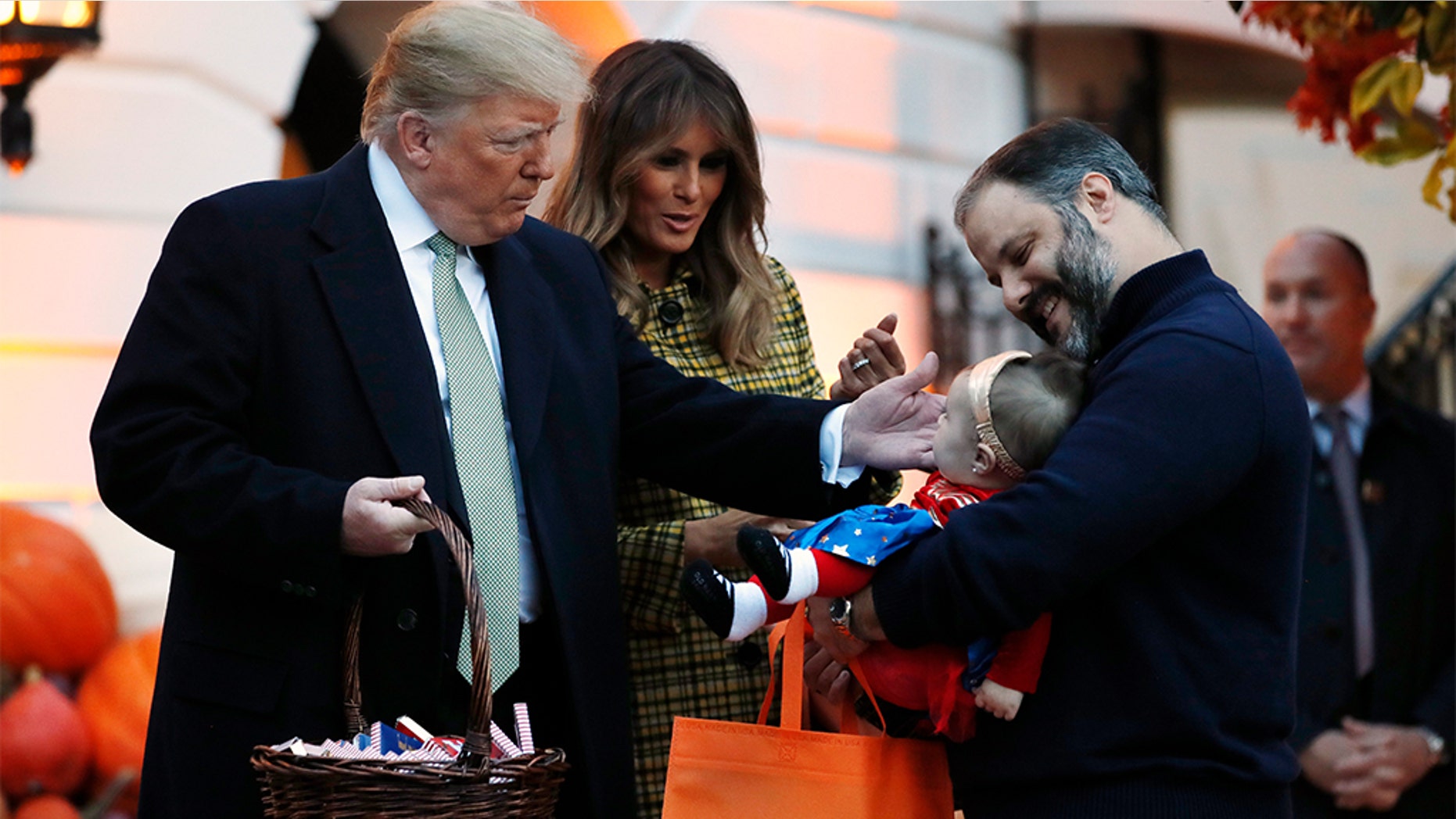 President Donald Trump and first lady Melania Trump greeting a baby in a superhero costume as they hand out candy to children during a Halloween trick-or-treat event at the White House.