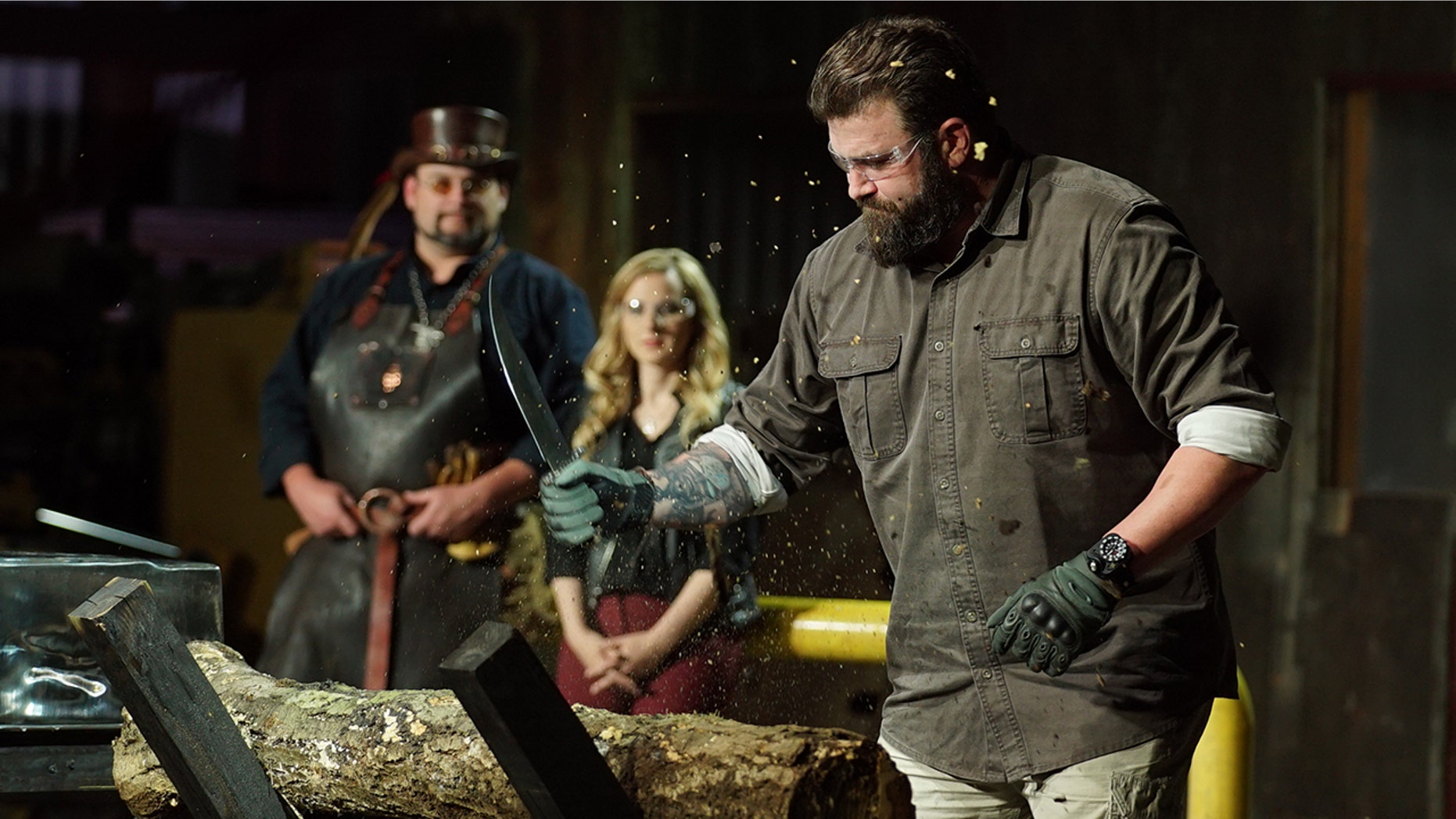'Master of Arms' judge Zeke chopping at a log with contestants knife. Trent and Ashley in the background.