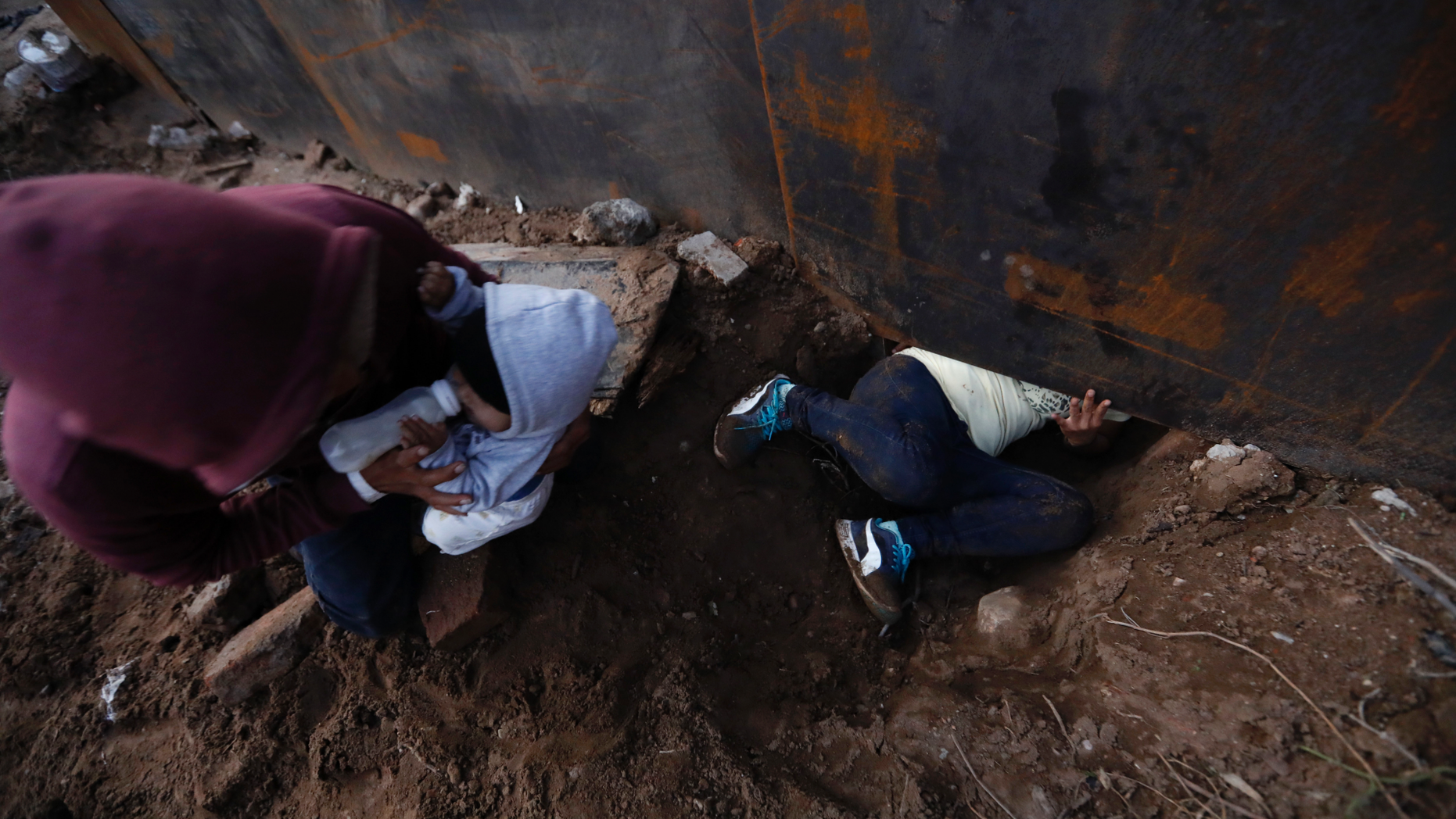 Honduran migrant Joel Mendez, 22, feeds his eight-month-old son Daniel as his partner Yesenia Martinez, 24, crawls through a hole under the U.S. border wall, in Tijuana, Mexico, Friday, Dec. 7, 2018. Moments later Martinez surrendered to waiting border guards while Mendez stayed behind in Tijuana to work, saying he feared he'd be deported if he crossed. (AP Photo/Rebecca Blackwell)
