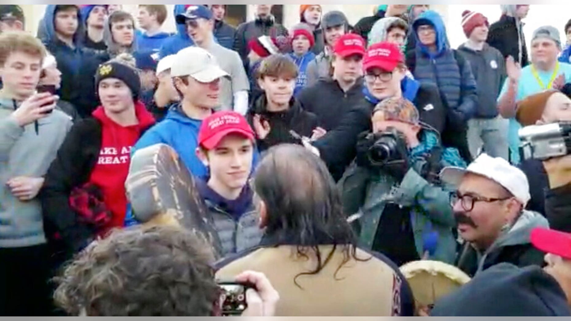 In this Friday, Jan. 18, 2019 image made from video provided by the Survival Media Agency, a teenager wearing a "Make America Great Again" hat, center left, stands in front of an elderly Native American singing and playing a drum in Washington. The Roman Catholic Diocese of Covington in Kentucky is looking into this and other videos that show youths, possibly from the diocese's all-male Covington Catholic High School, mocking Native Americans at a rally in Washington. (Survival Media Agency via AP)