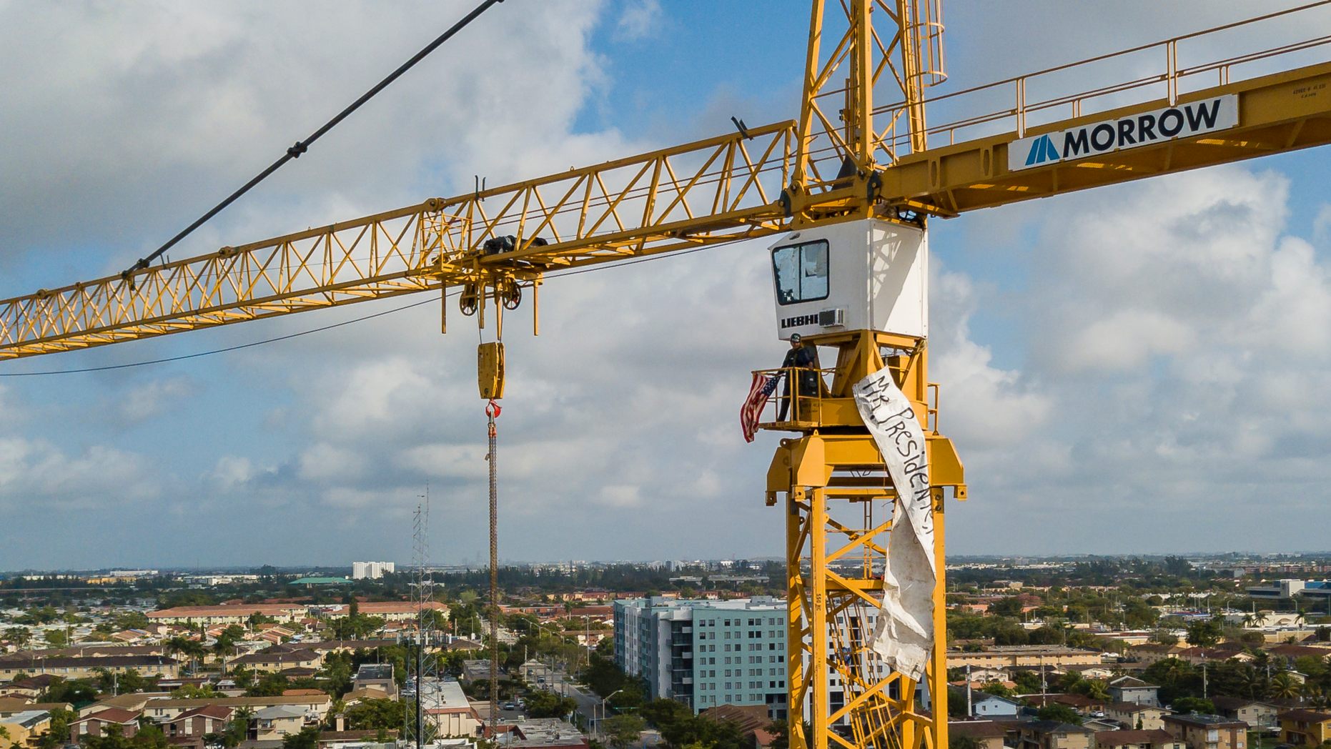 A man stands inside a construction crane where he secured an American flag and banner near Florida International University in Sweetwater, Florida on Monday, February 18, 2019. President Donald Trump must organize a meeting at school in the afternoon. (Matias J. Ocner / Miami Herald via AP)