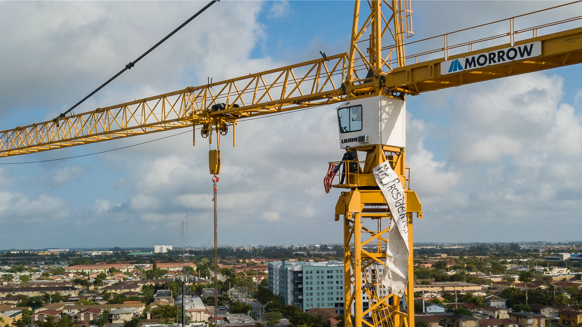 A man stands inside a construction crane where he attached an American flag and a banner near Florida International University in Sweetwater, Fla., Monday, Feb. 18, 2019. President Donald Trump is scheduled to host a rally at the school in the afternoon. (Matias J. Ocner/Miami Herald via AP)