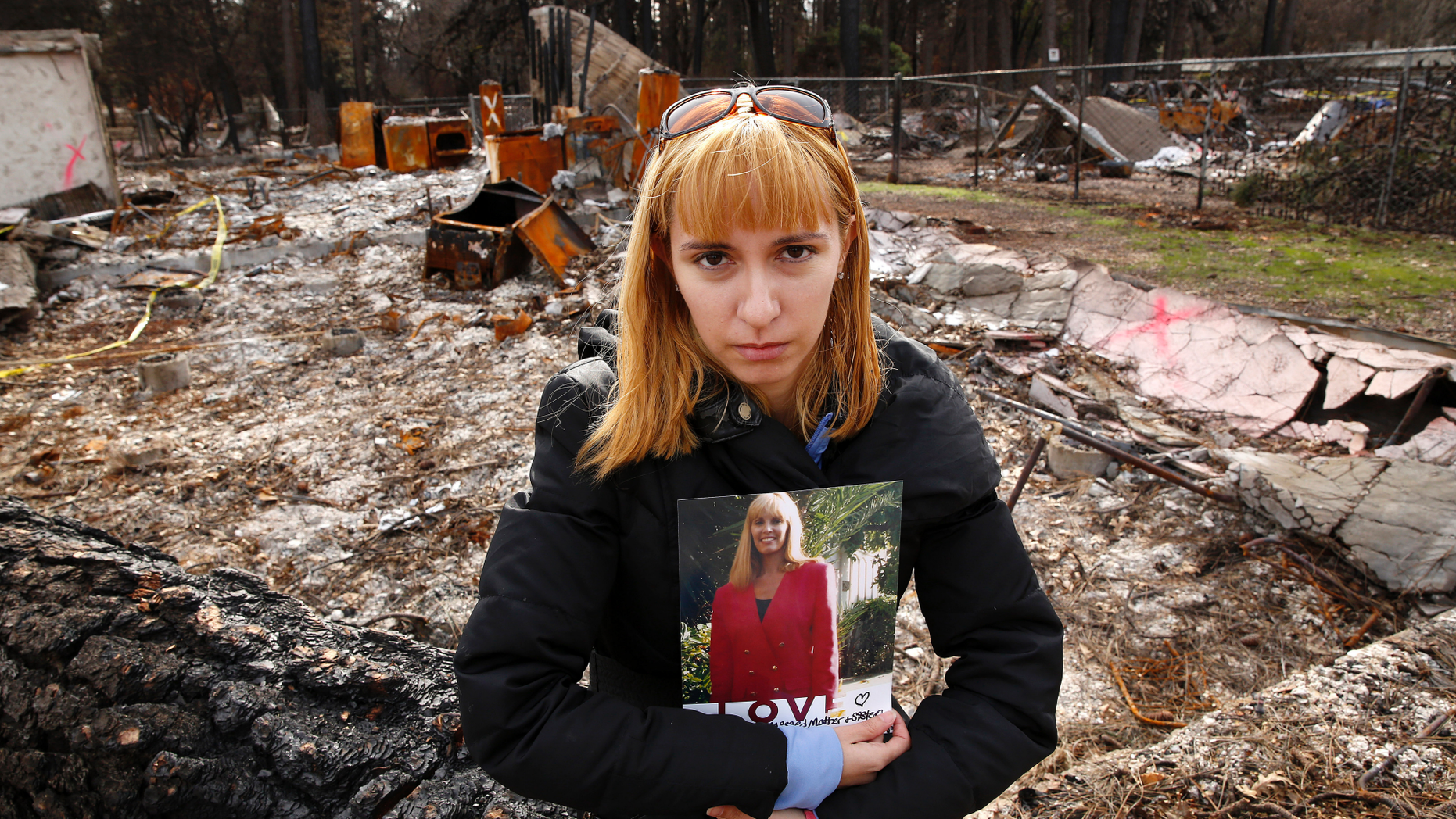 In this Thursday, Feb. 7, 2019 photo, Christina Taft, the daughter of Camp Fire victim Victoria Taft, poses with a photo of her mother, at the burned out ruins of the Paradise, Calif., home where she died last fall. Taft refused to leave. If the threat was real, authorities would order an evacuation, she told her daughter. Victoria Taft’s remains were recovered from the ruins of her living room. (AP Photo/Rich Pedroncelli)