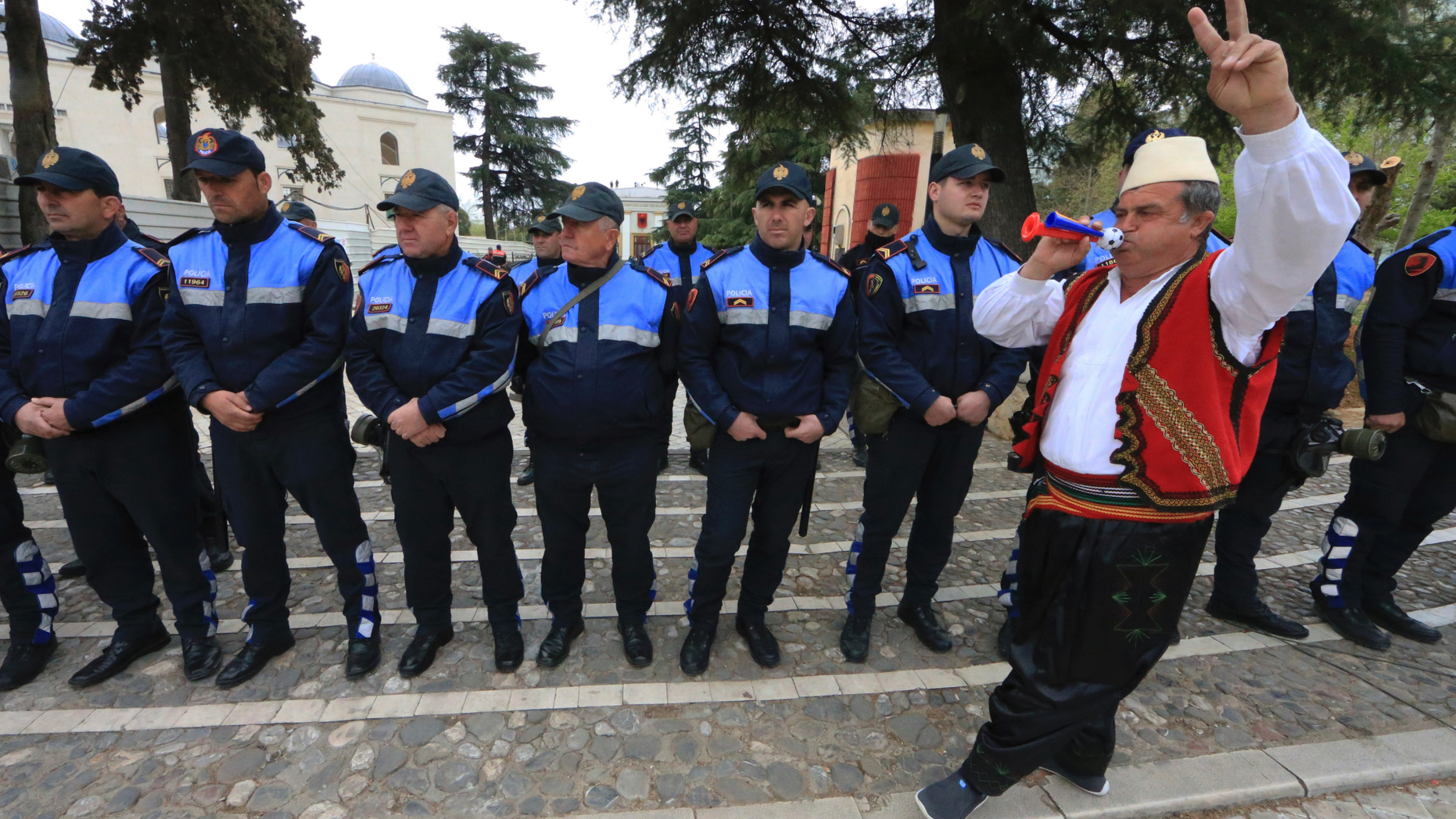A protester dressed in traditional clothing flashes the victory sign in front of Albanian policemen guarding the parliament building in Tirana, on Thursday, March 28, 2019. Albanian opposition protesters have repeated attempts to enter the parliament by force in their protest asking for the government's resignation and an early election. (AP Photo/ Hektor Pustina)