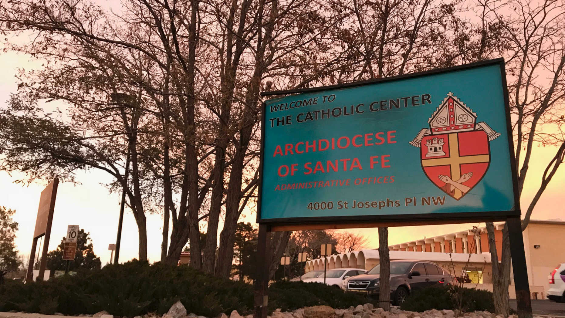 FILE - In this Nov. 29, 2018, file photo, the sun sets on a sign in front of the Archdiocese of Santa Fe offices in Albuquerque, N.M. A priest who fled the United States decades ago faces a federal trial on charges that he sexually abused a New Mexico boy in the early 1990s at an Air Force base and veterans' cemetery. Arthur Perrault, a one-time pastor in Albuquerque, has been charged with aggravated sexual abuse and others counts. (AP Photo/Susan Montoya Bryan, File)