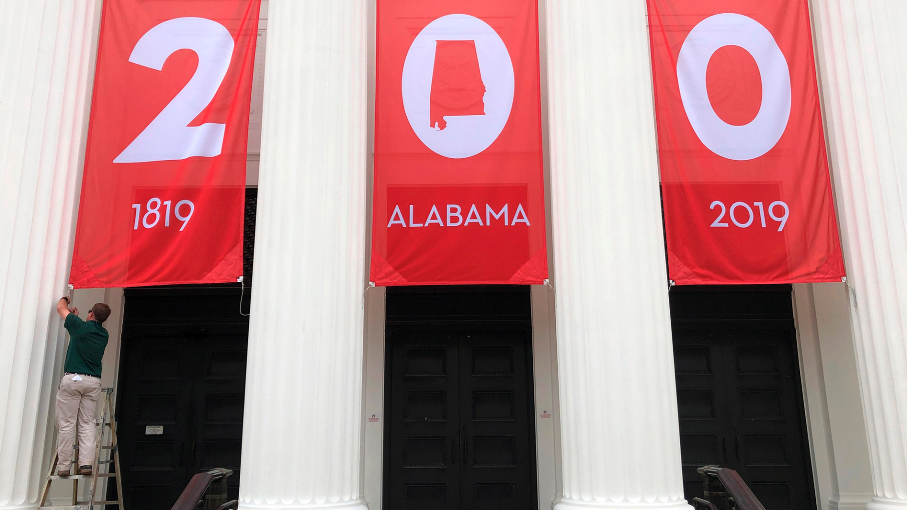In this Feb. 28, 2019, photo a worker adjusts a banner celebrating Alabama's bicentennial outside the Department of Archives and History in Montgomery, Ala. Alabama is the latest state trying to balance a painful past with other parts of its history as it recognizes its 200th birthday as a state. (AP Photo/Jay Reeves)