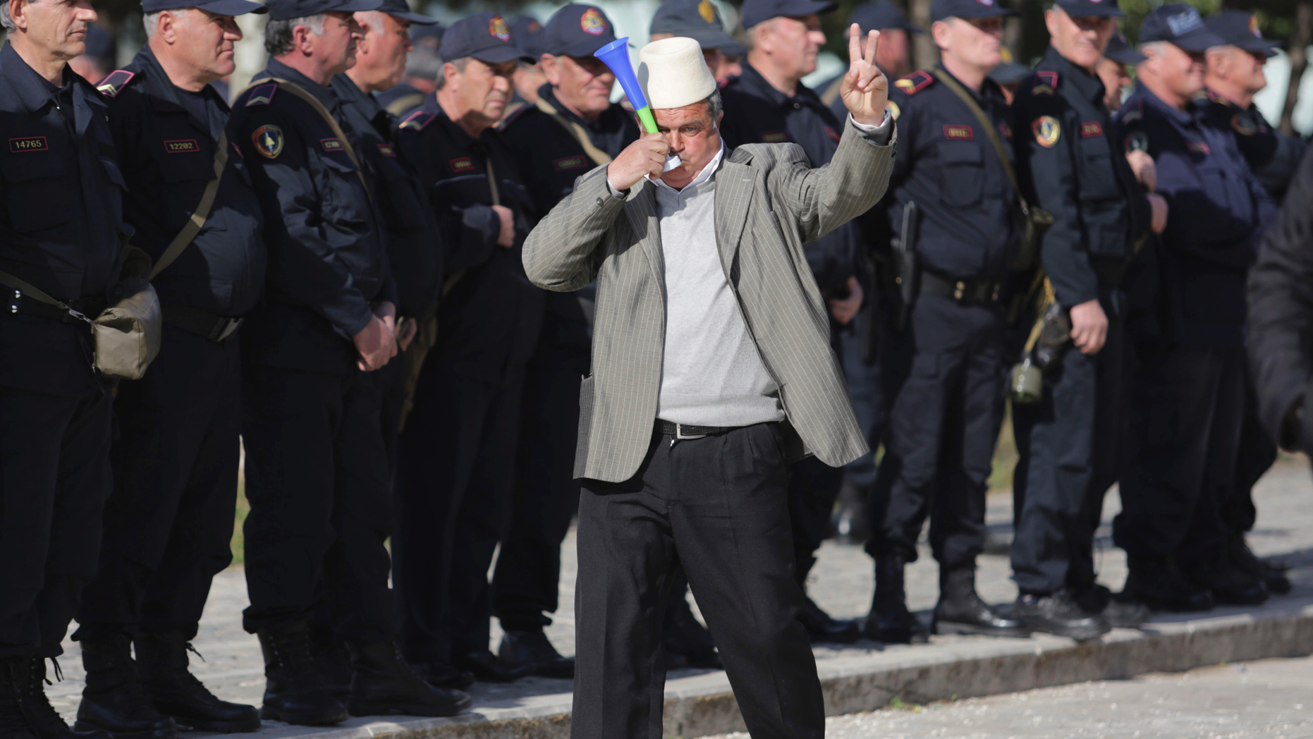 A protester flashes the victory sign in front of Albanian policemen guarding the parliament building as opposition supporters gather to take part in an anti-government rally , in Tirana , Albania, Tuesday, March 5, 2019. The opposition demanded the government step down, claiming it's corrupt and has links to organized crime. (AP Photo/ Hektor Pustina)