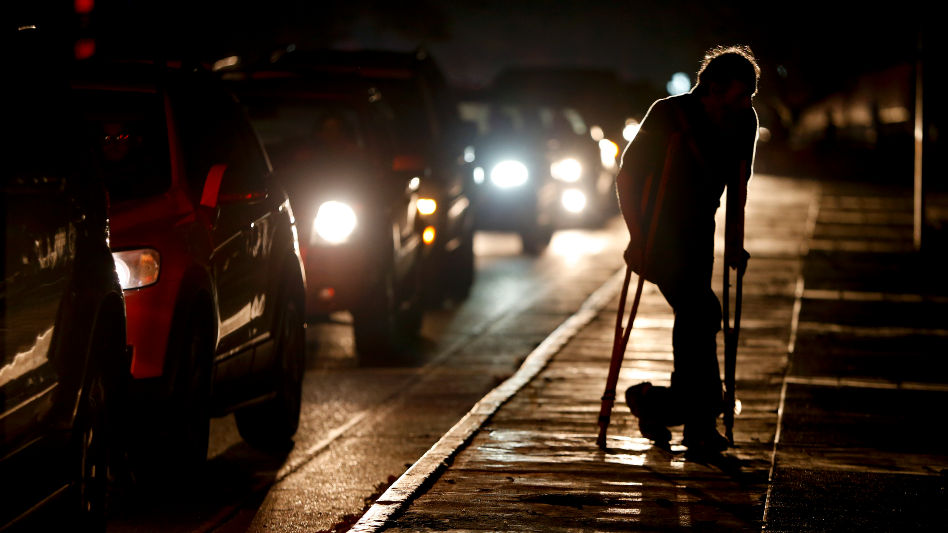FILE - In this March 29, 2019 file photo, a man on crutches is illuminated by headlights of oncoming traffic, in Caracas, Venezuela. Venezuelans are struggling to understand the Sunday, March 31, 2019 announcement that the nation’s electricity is being rationed to combat daily blackouts.  (AP Photo/Natacha Pisarenko, File)