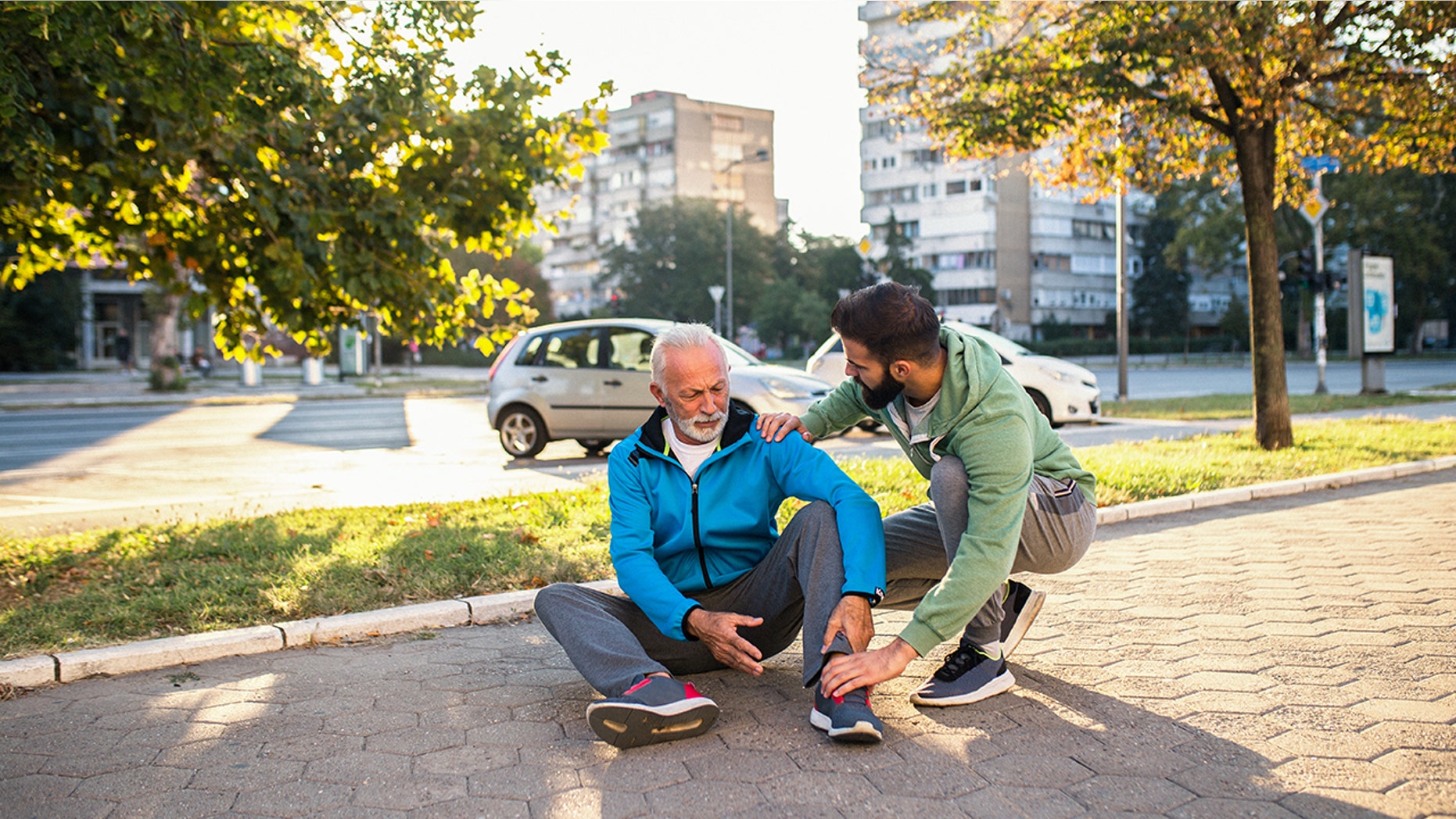 Son helping his father to stand up after his fall while jogging outdoors in city street.